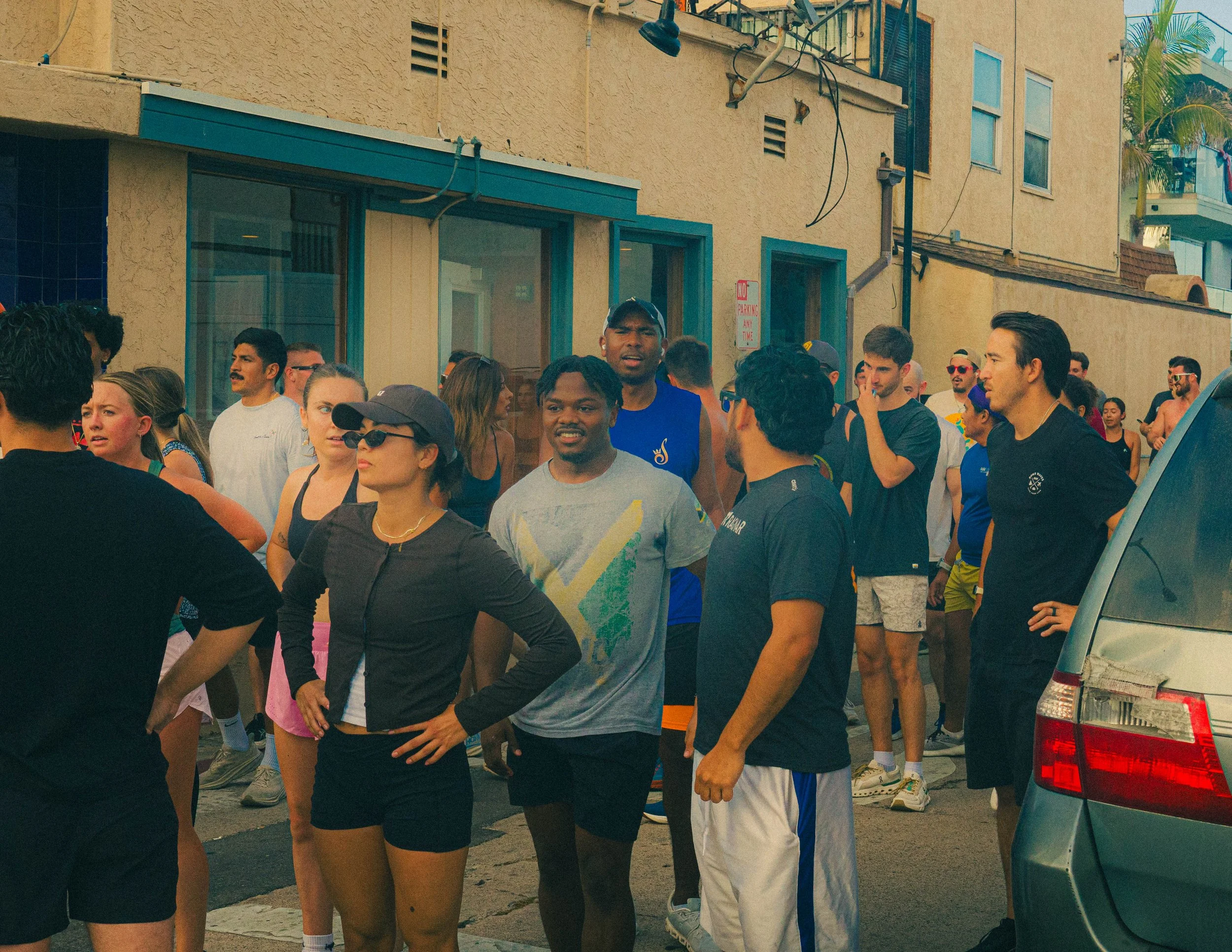 A group of people gathered outdoors on a city street, with some engaged in conversation and others standing around, some wearing athletic clothing and sunglasses.