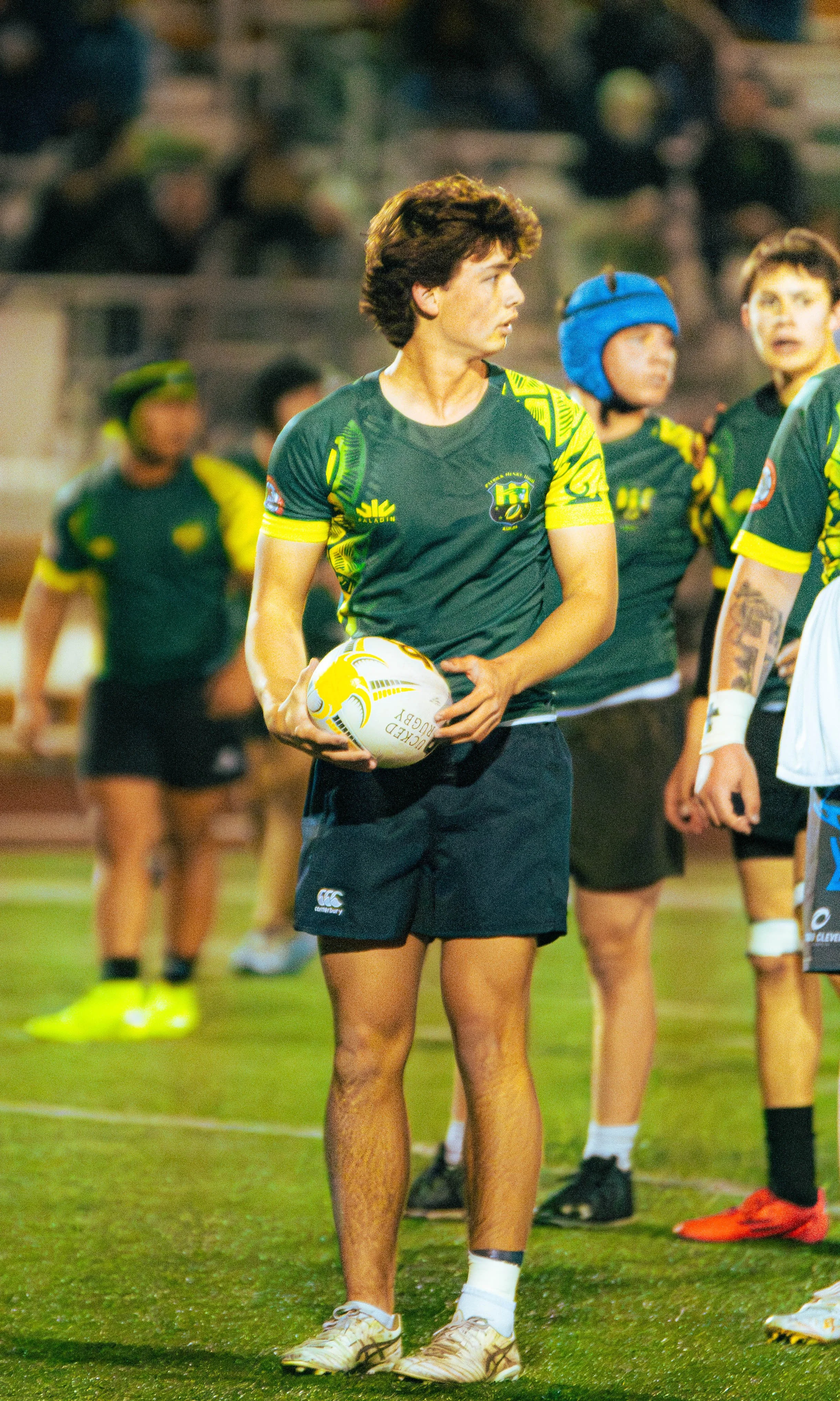 A rugby player in a green and yellow uniform holding a rugby ball, standing on a field with teammates and spectators in the background.