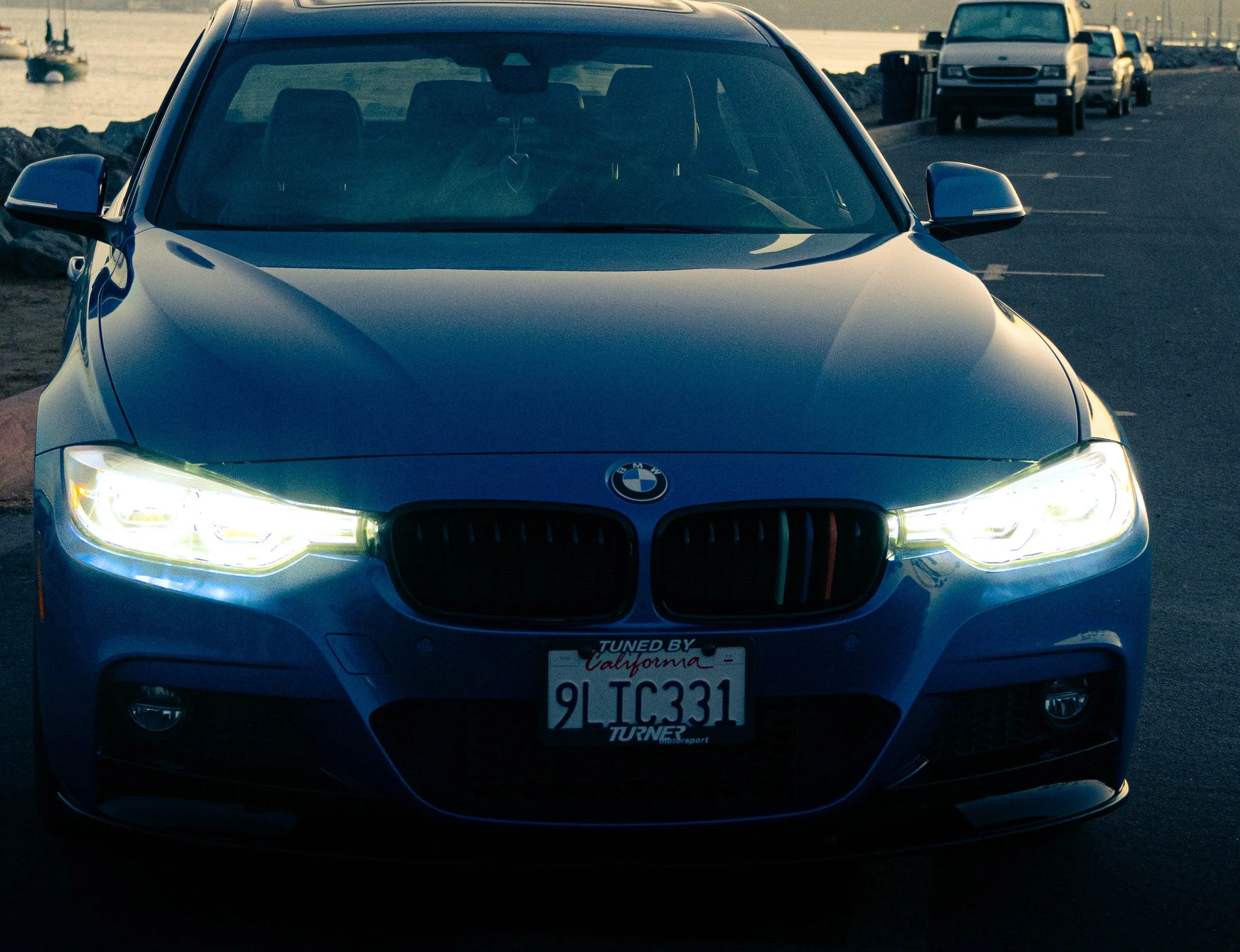 Front view of a silver BMW car parked near the water with other vehicles and a boat in the background during sunset or sunrise.