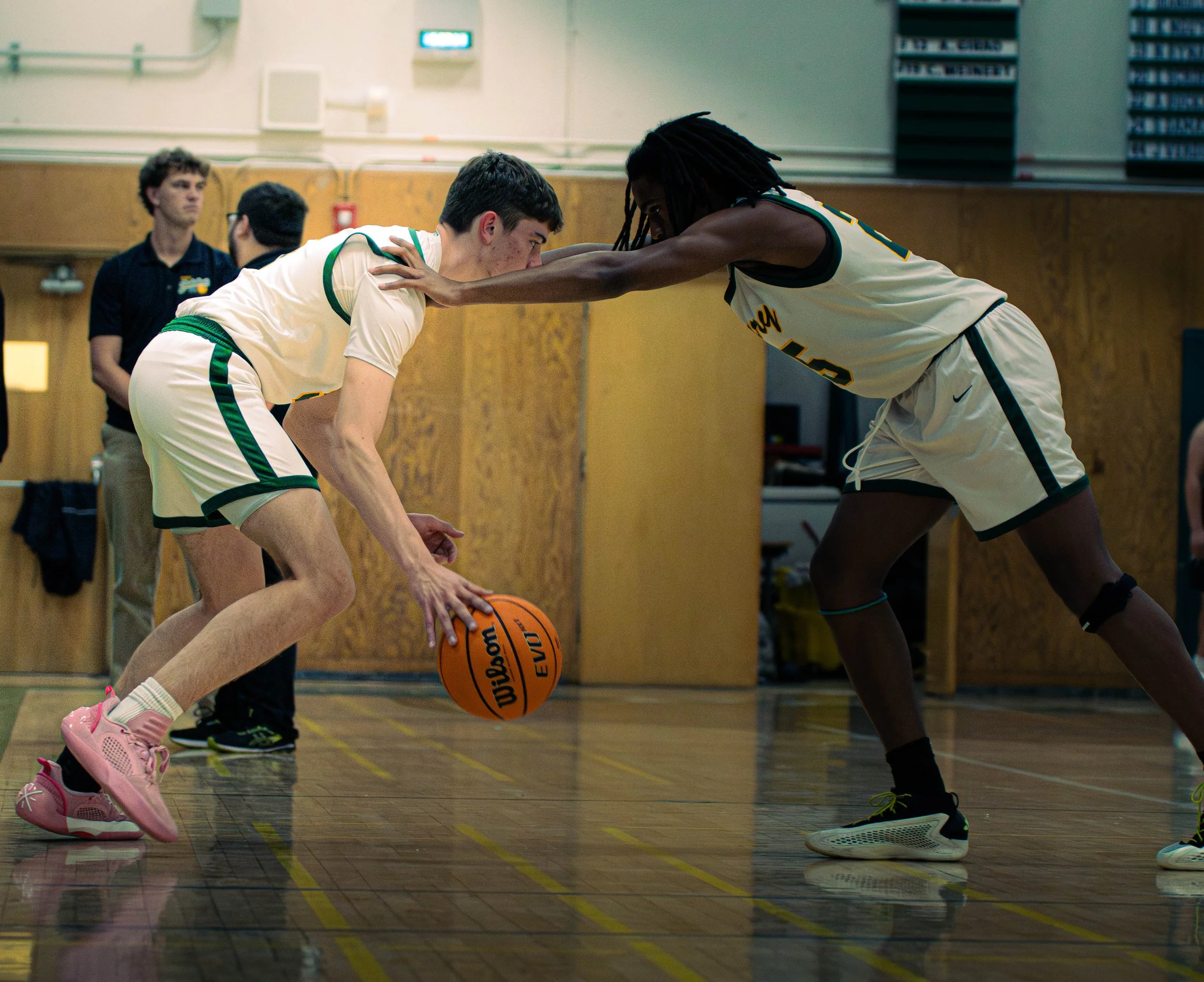 Two basketball players, one in a white jersey and the other in a black and white jersey, are competing closely on a gymnasium court as they reach for a basketball. There are a few people in the background watching, including a young man wearing glass