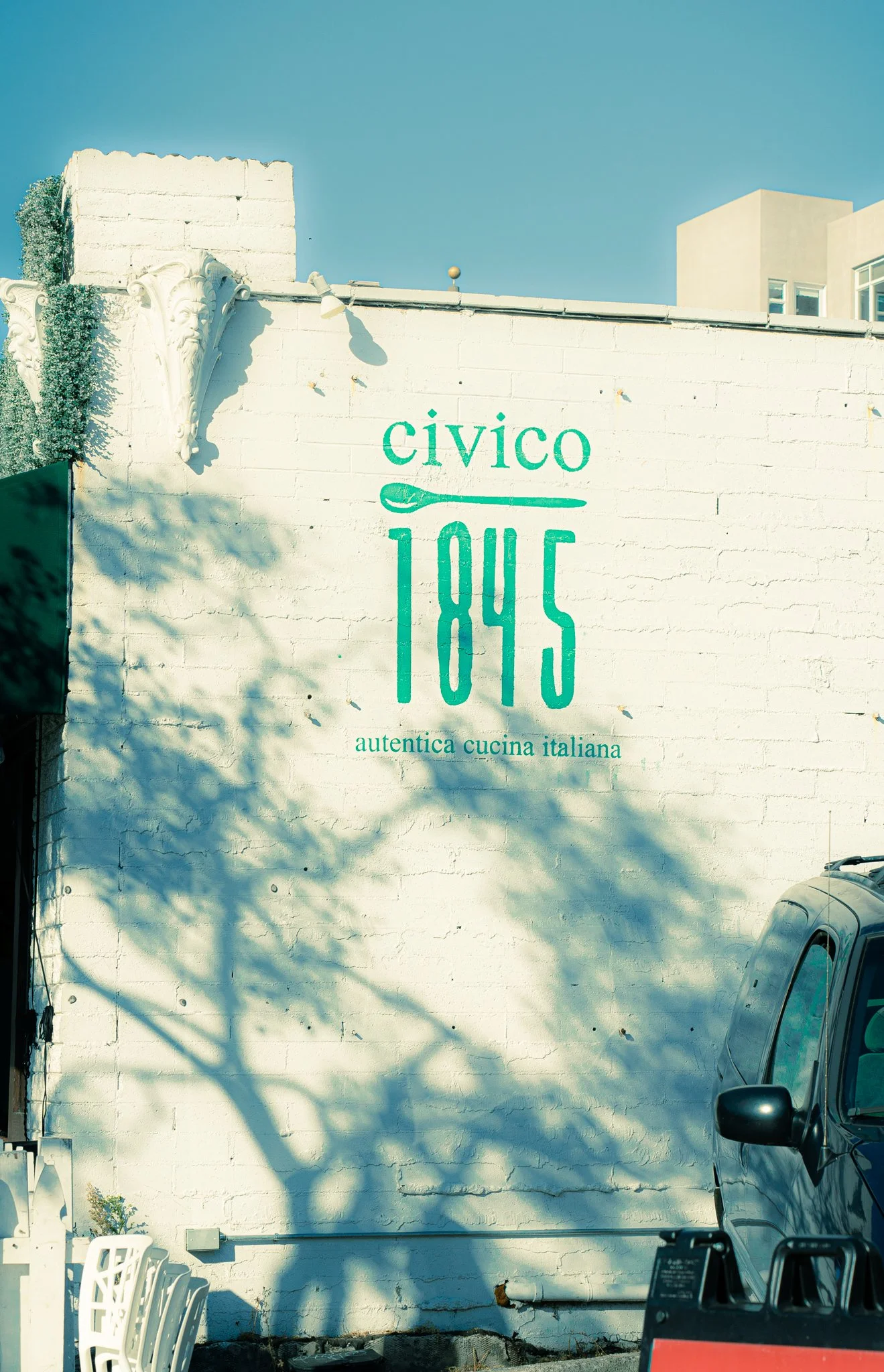 A white brick wall with green text that reads "civico 1845" and below that "autentica cucina italiana." There are chairs in the bottom left corner and a vehicle on the right side, with shadows cast on the wall from nearby trees against a clear blue s