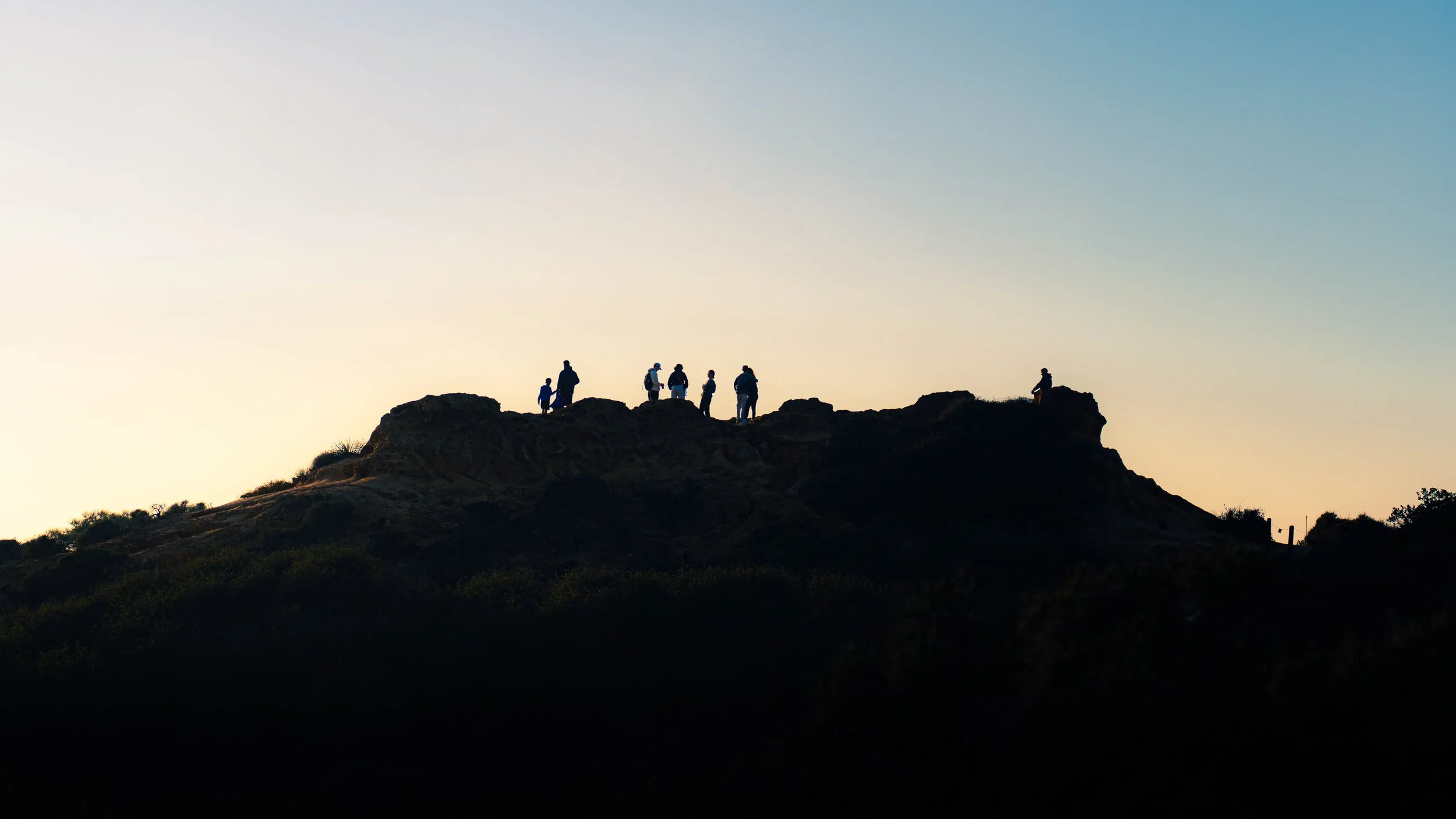 Silhouettes of people standing on a rocky hilltop at sunset or sunrise, with a clear sky in the background.