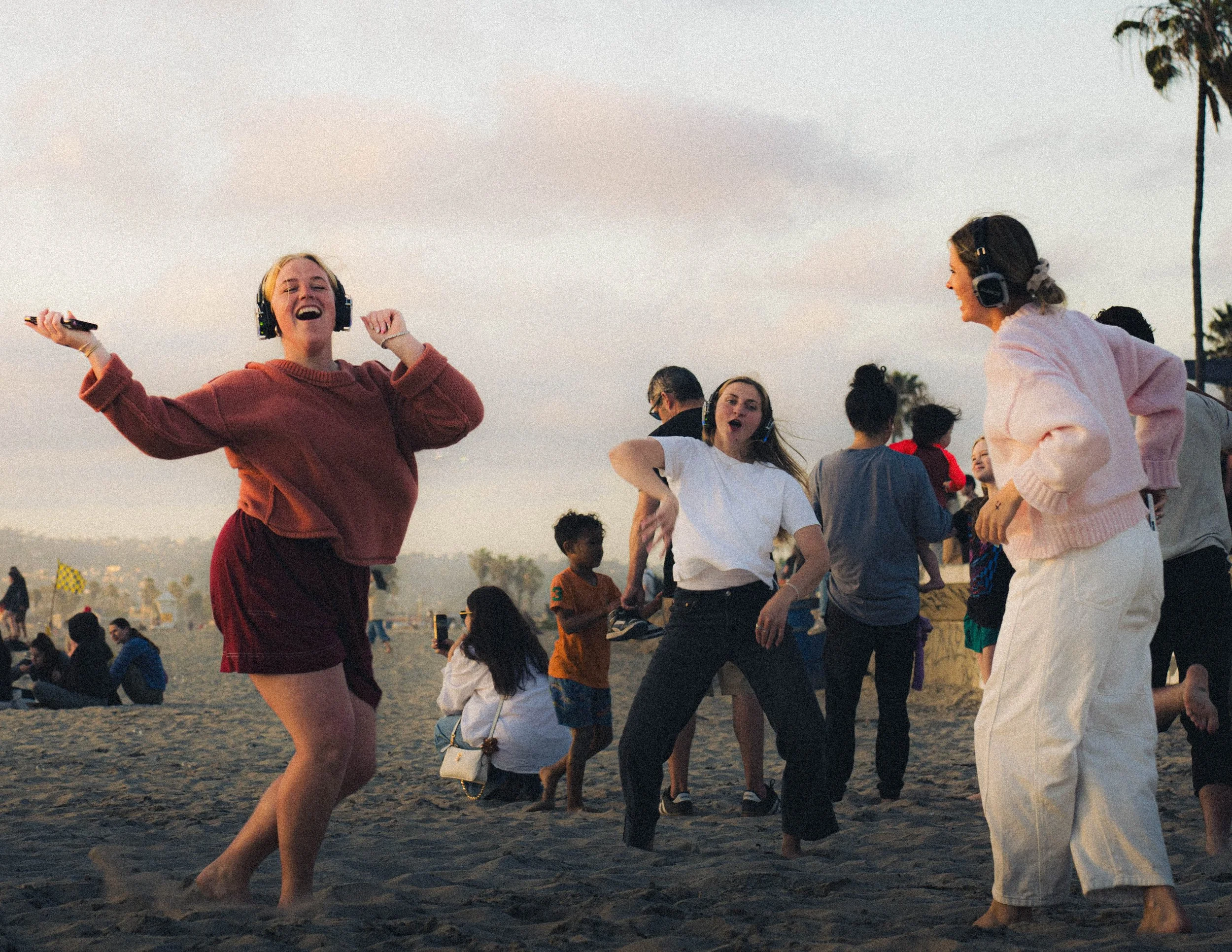 People dancing and enjoying music at a beach during sunset, with palm trees in the background.