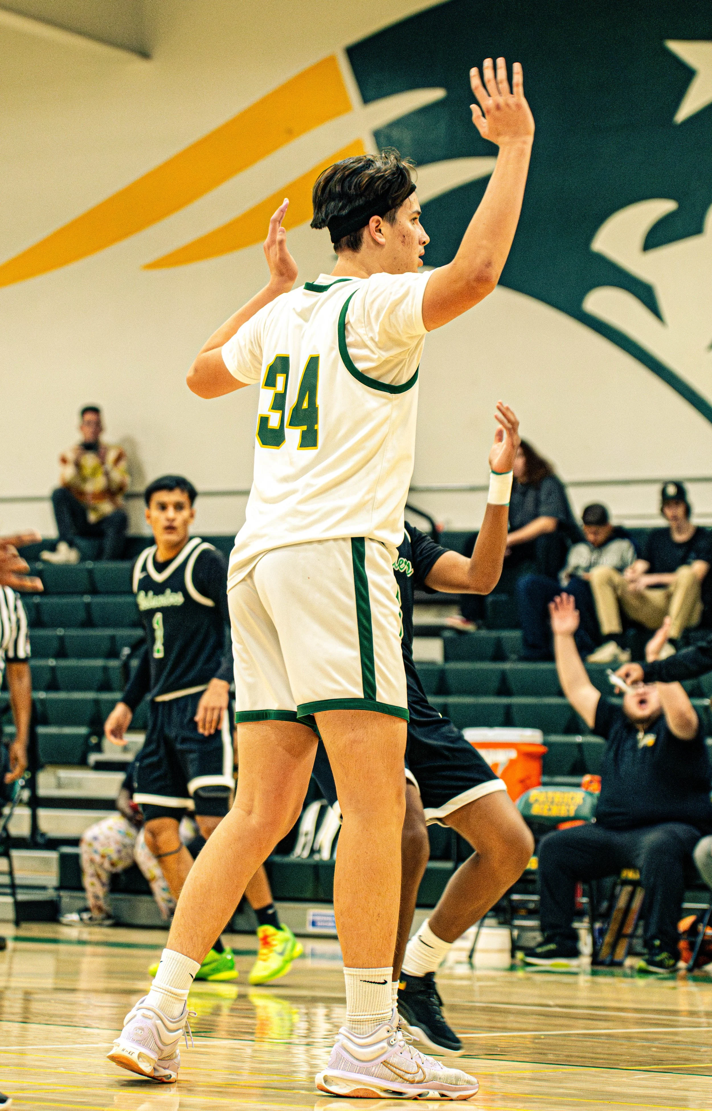 A female volleyball player in a white uniform with green and yellow accents, wearing Nike sneakers, during a game or practice in an indoor gym, with spectators and other players in the background.