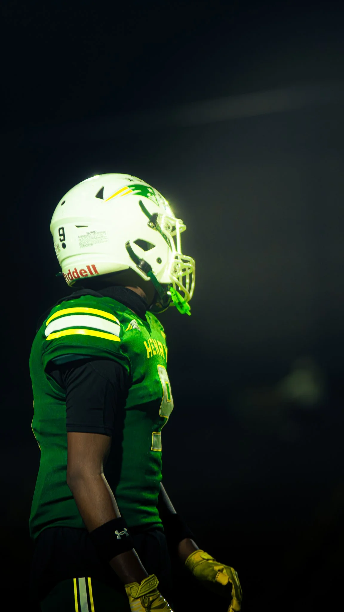 A football player in a green uniform with yellow accents, wearing a white helmet with a green and yellow logo, standing against a dark background.