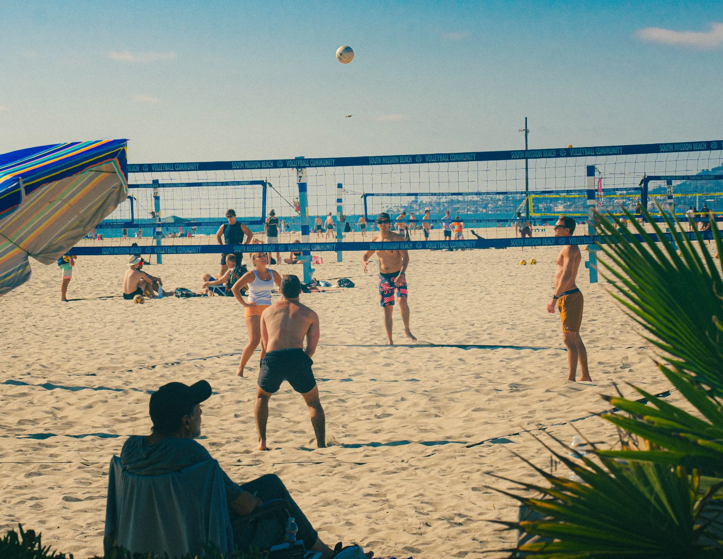 People playing beach volleyball on a sandy court at South Mission Beach with a hot air balloon and blue sky in the background.