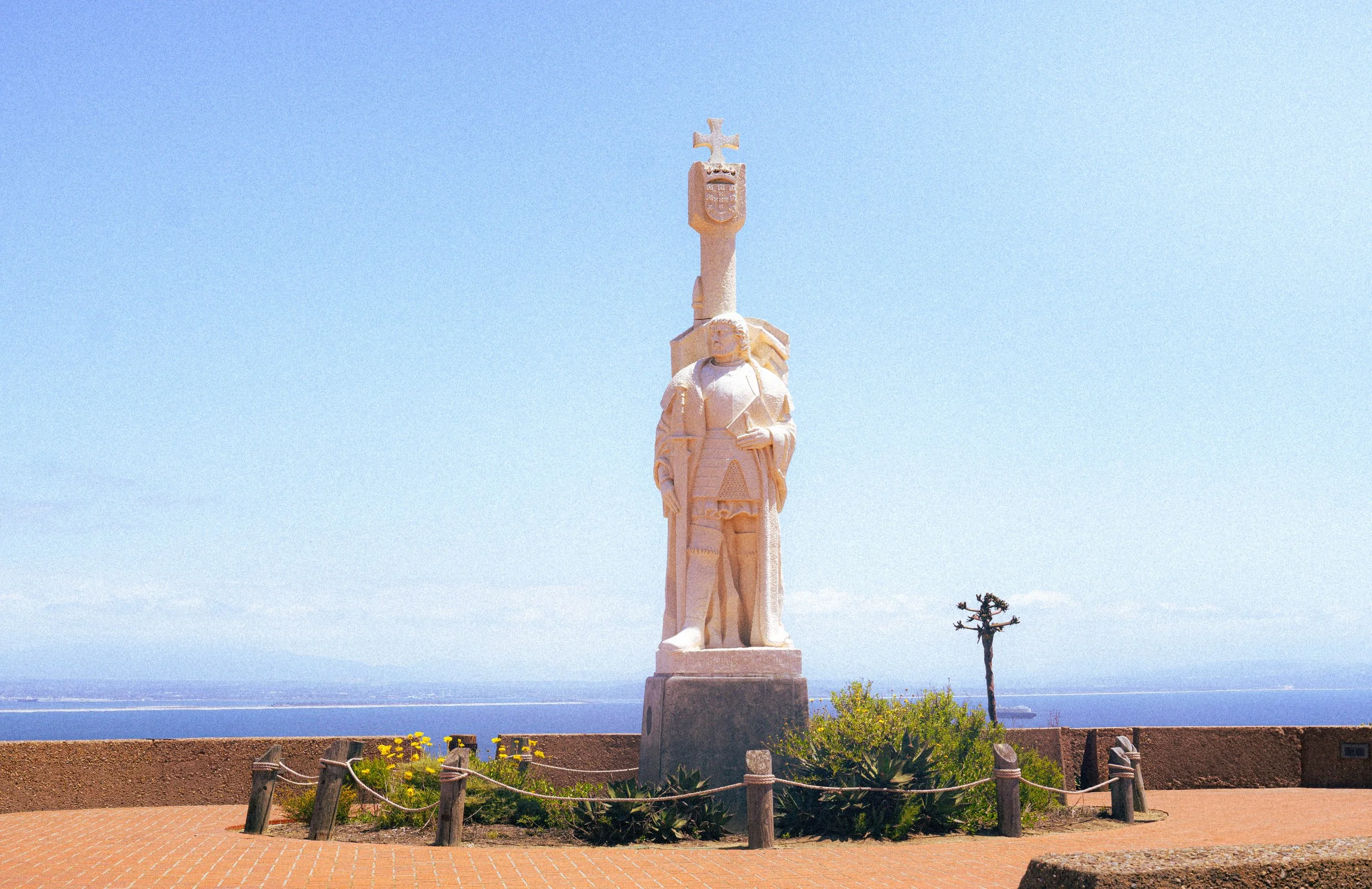 Statue of a historic figure holding a book, topped with a cross and shield emblem, against a clear blue sky, near the ocean in a park with flowers and a small tree.