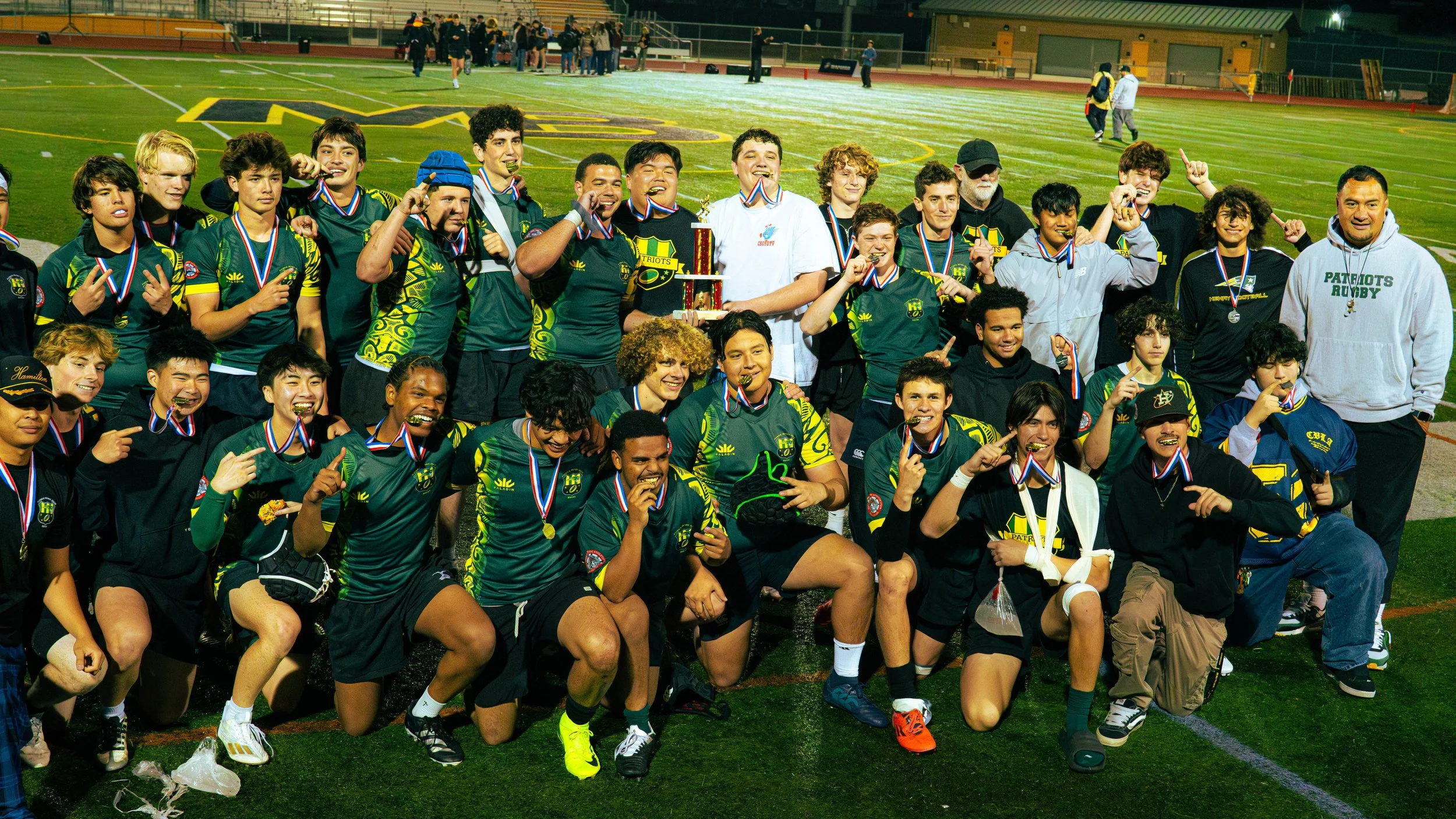A group of young athletes, some wearing green and yellow uniforms, celebrating on a sports field with medals around their necks and a trophy, indicating they won a sports event. Some are making victory signs, and they are posing for a team photo.