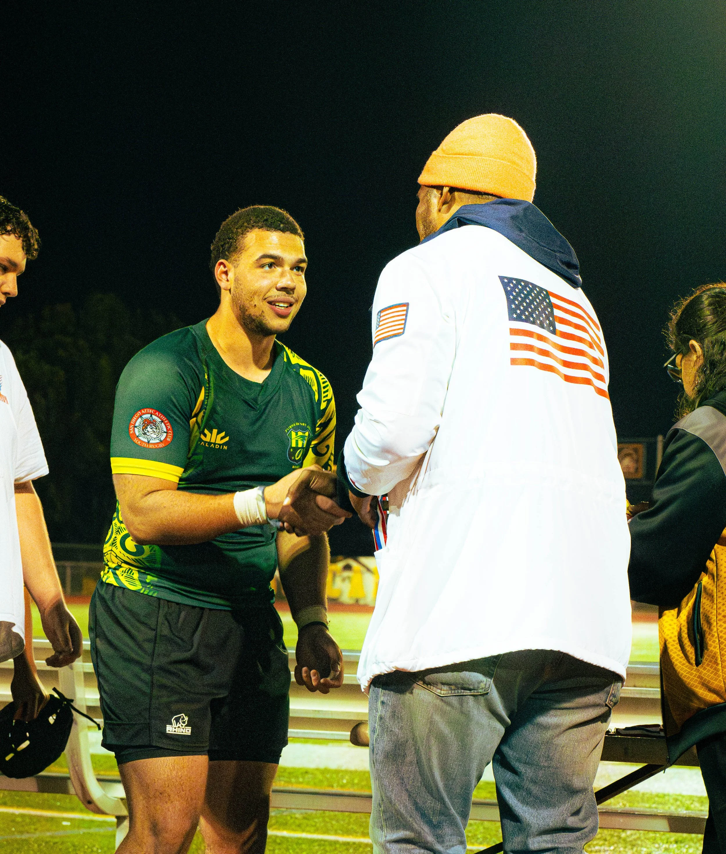 Group of young men on a sports field during nighttime, engaged in conversation, with a man wearing a white jacket with an American flag and orange beanie. Others wear sports jerseys and casual attire.