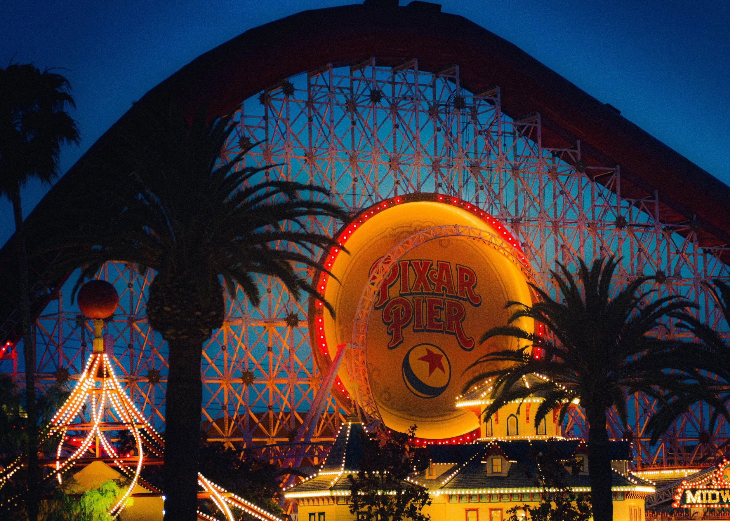 Nighttime view of Pixar Pier at Disney California Adventure Park, featuring a large illuminated Pixar ball sign with the park's name, surrounded by colorful lights, palm trees, and part of a roller coaster structure.
