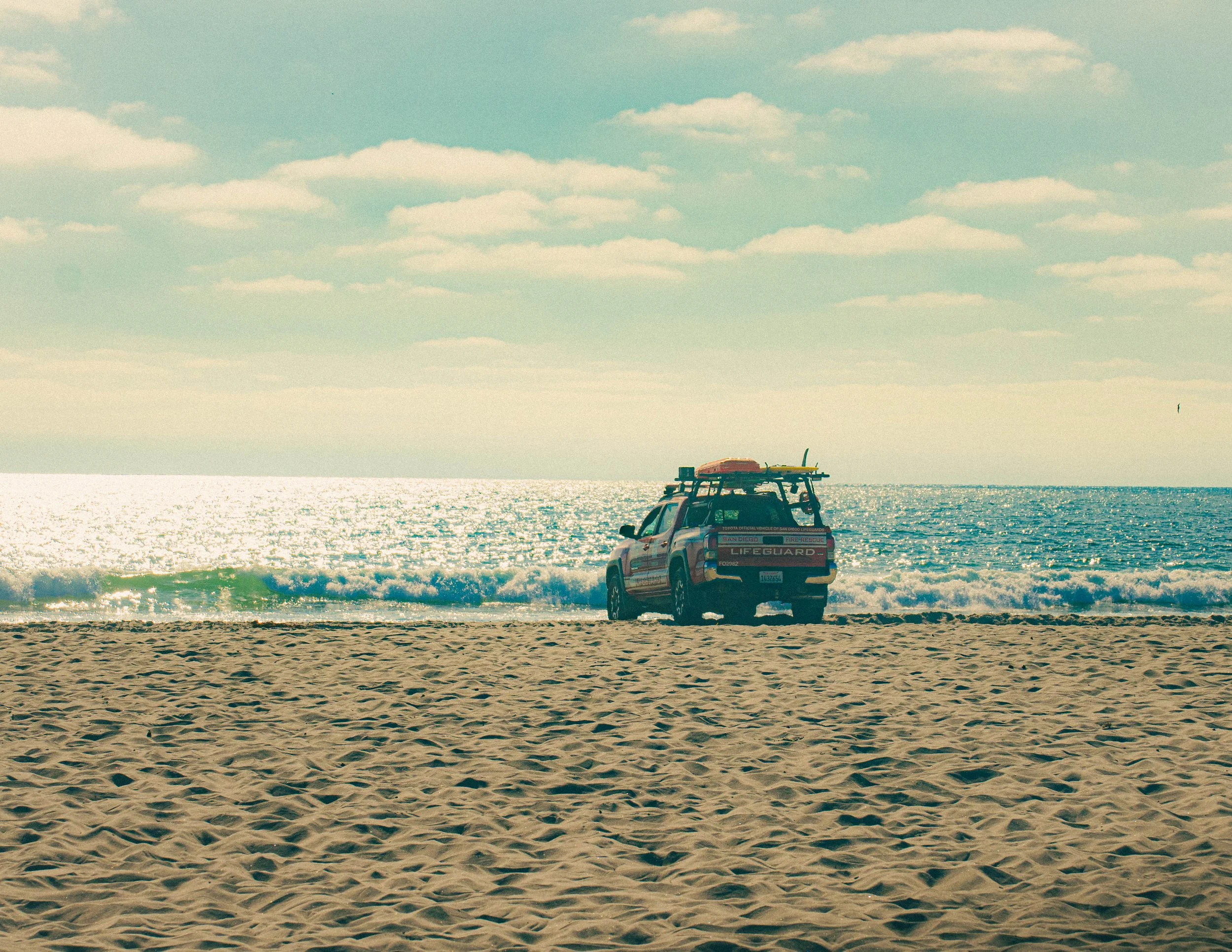 A lifeguard vehicle with a surfboard on top is parked on a sandy beach, with the ocean and a partly cloudy sky in the background.
