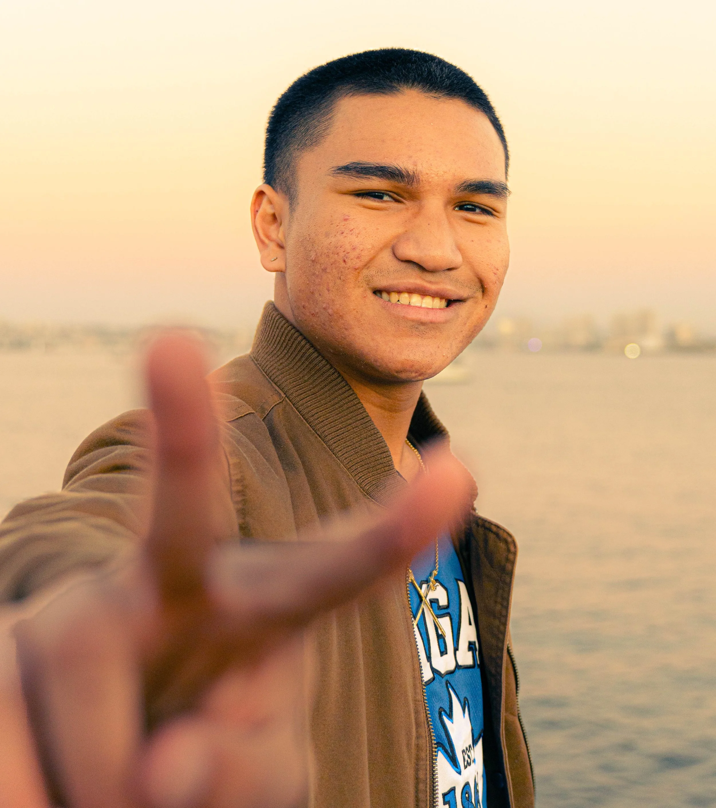 Close-up of a young man with short black hair smiling at the camera, blurred background of water and sky, wearing a brown jacket and a blue shirt with white and black text, with a finger partially blocking the camera lens.