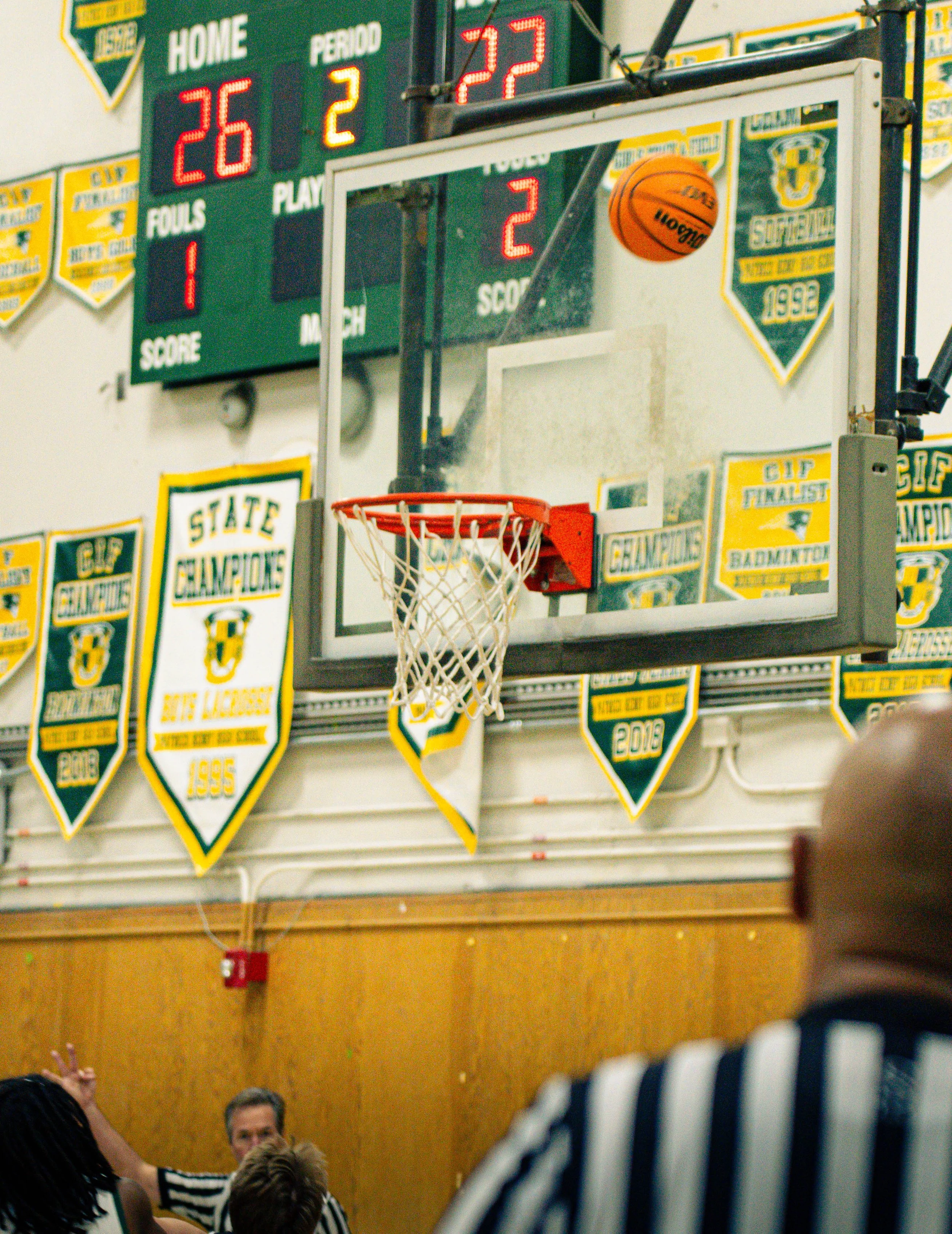 A basketball about to go through a basketball hoop during a game in a gymnasium, with a scoreboard in the background showing 26 home team points, 2 period, 22 away team points, and 1 foul.