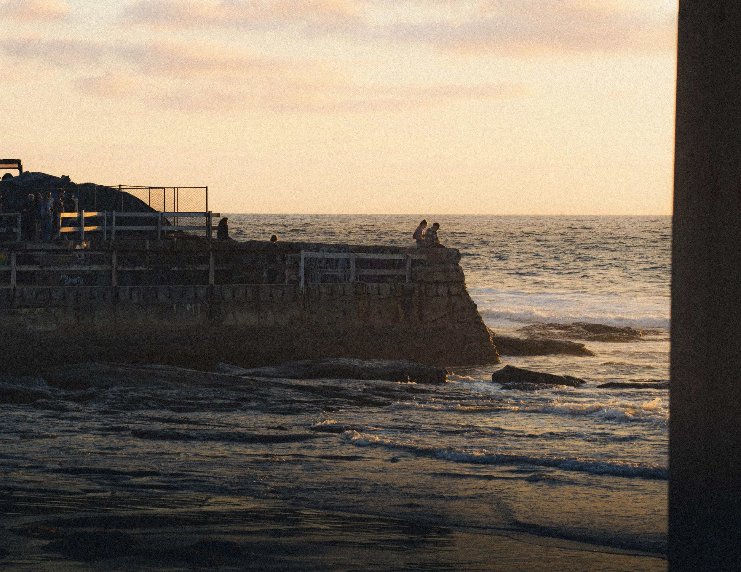 People standing and sitting on a pier by the ocean during sunset, with waves and a stone wall in the foreground.