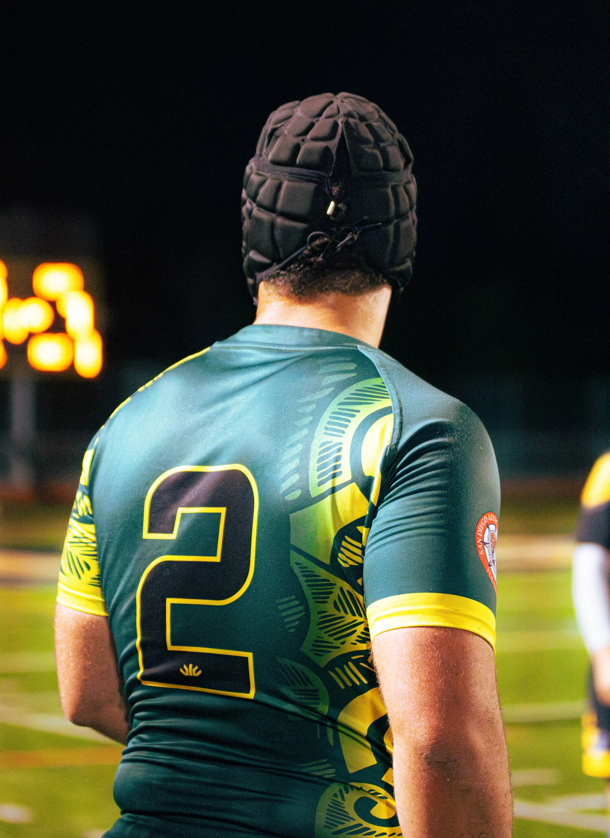 Back of a person wearing a green and yellow sports jersey with the number 2, standing on a sports field at night.