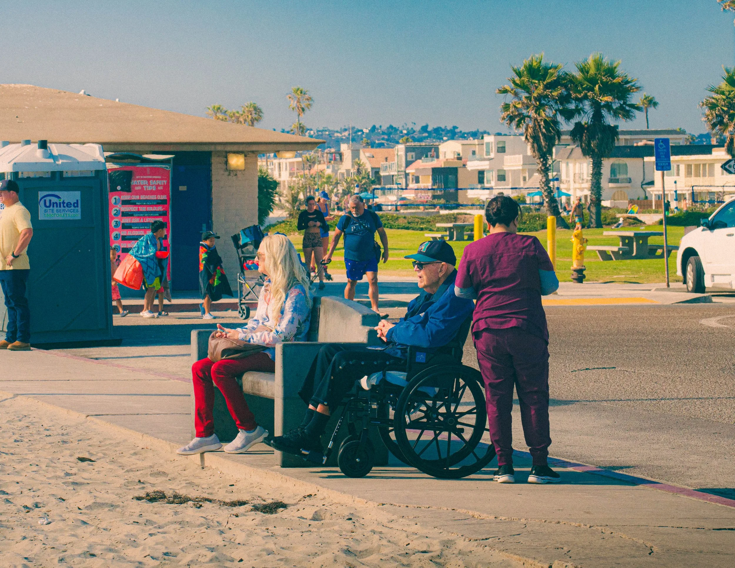 People sitting on benches and standing near a beachside pavilion with palm trees and buildings in the background on a sunny day.