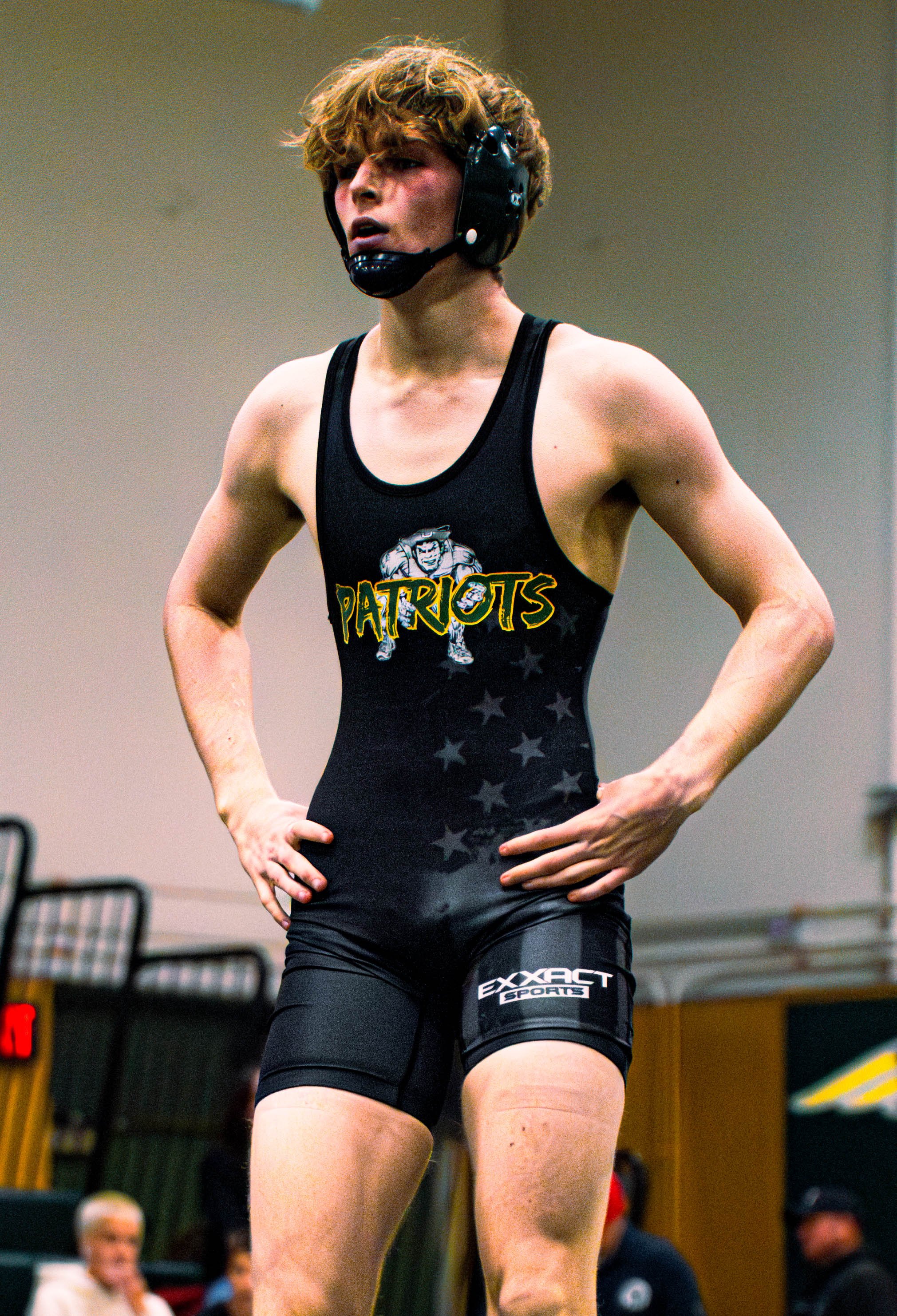 A female athlete with curly blonde hair wearing a black wrestling singlet with 'Patriots' logo and a black helmet, standing with hands on hips in an indoor sports facility. Spectators are blurred in the background.