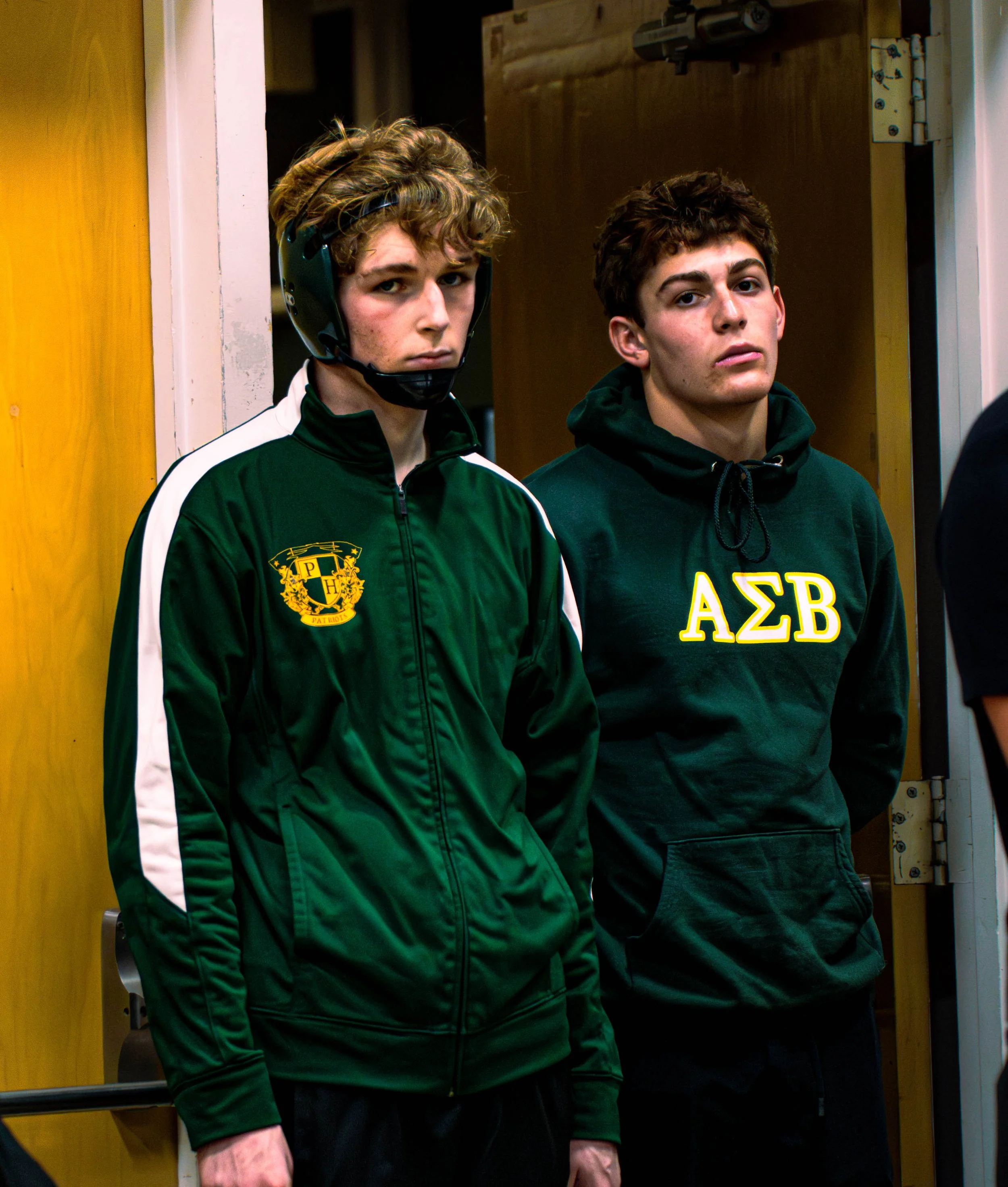 Two young men standing side by side in a doorway, wearing dark athletic jackets, one with a helmet and the other with the letters 'ΑΣΒ' on their sweatshirts, looking seriously at the camera.