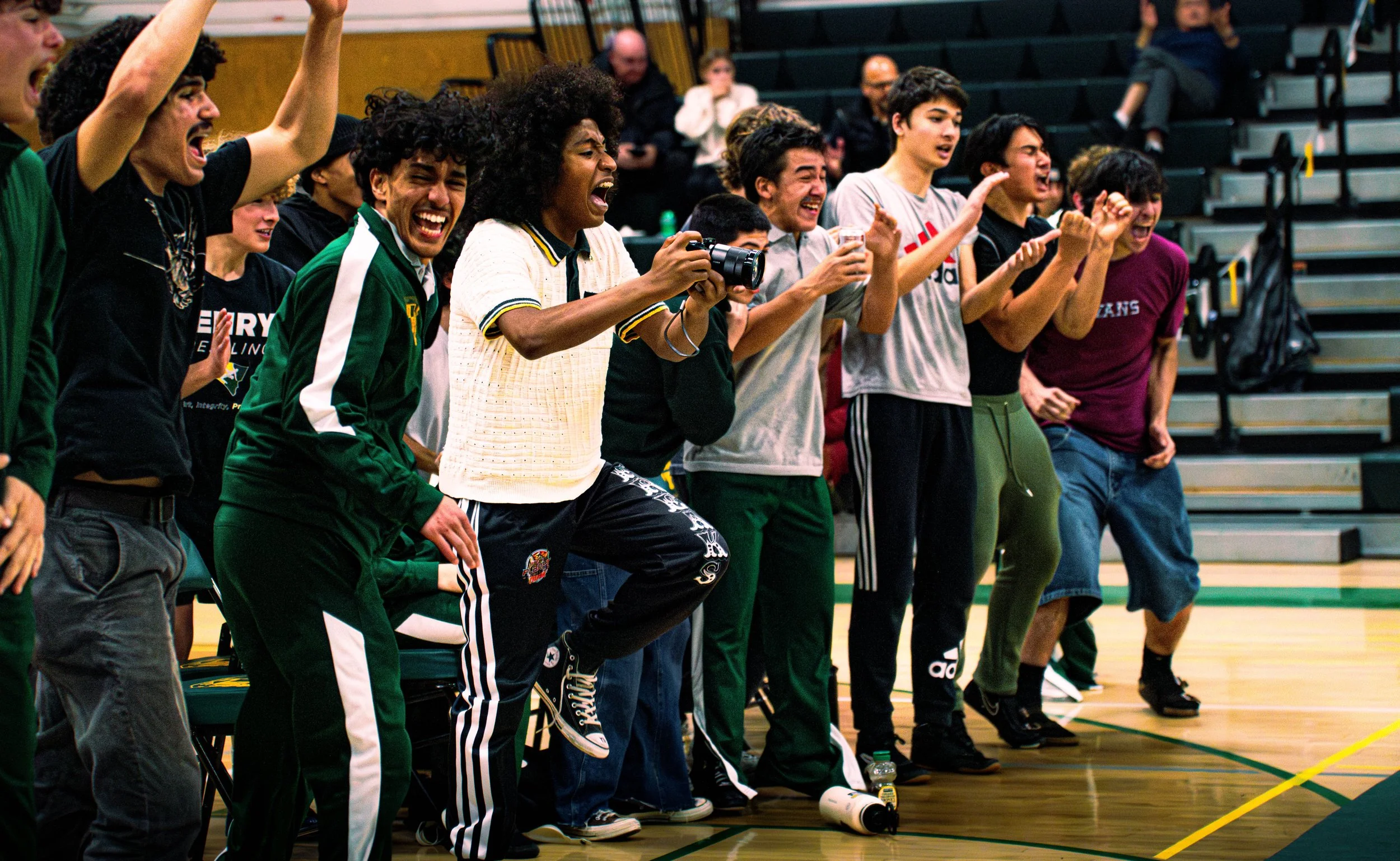 A group of young people, likely sports fans, cheering and celebrating from the sidelines of an indoor sports event in a gymnasium. They appear excited, some with open mouths and fists raised.