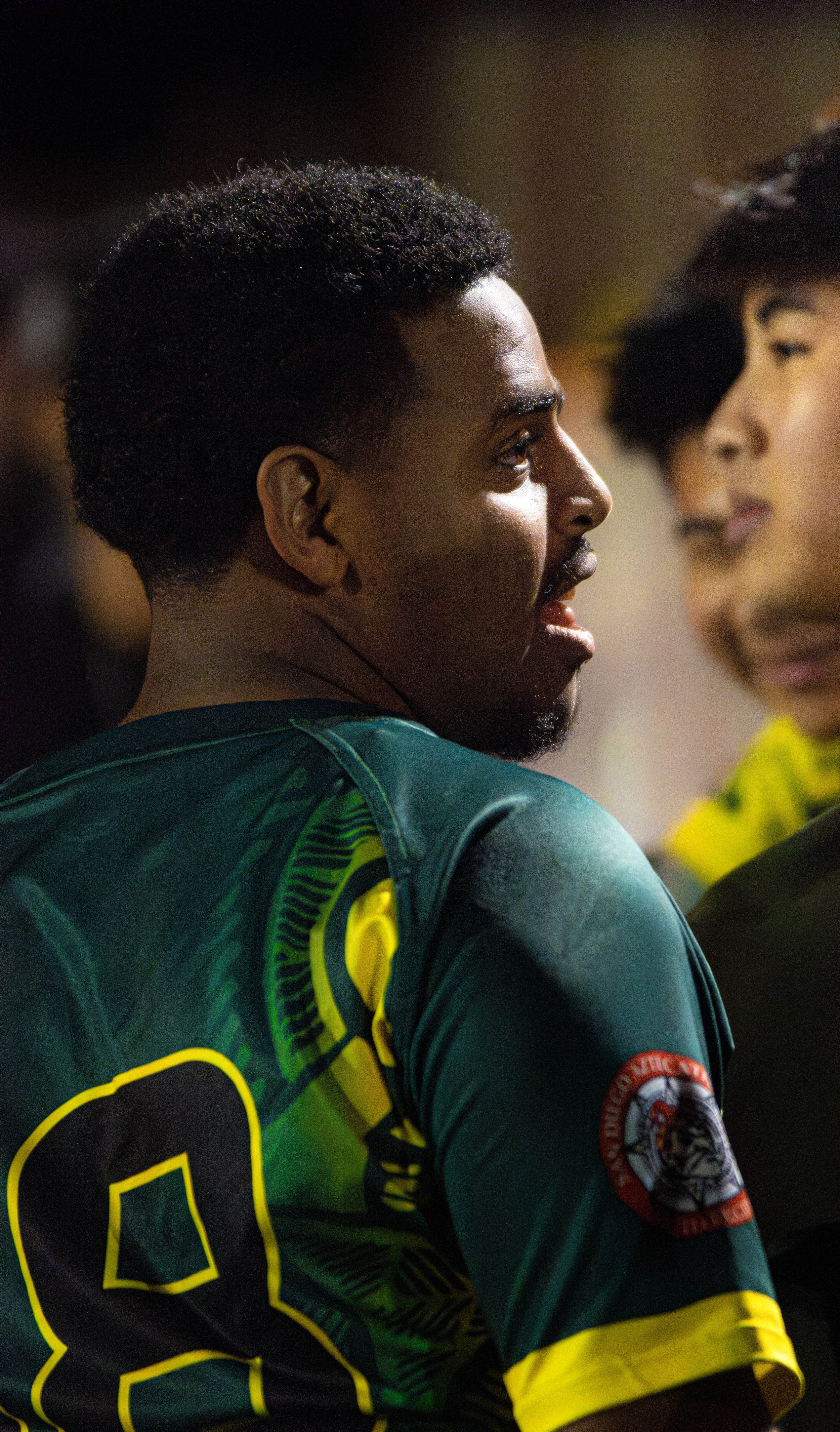 A man with dark skin and short curly hair, facing right, wearing a dark green sports jersey with yellow accents and a badge on the sleeve, and a woman with dark hair and lighter skin, also facing right, partially visible in the background.