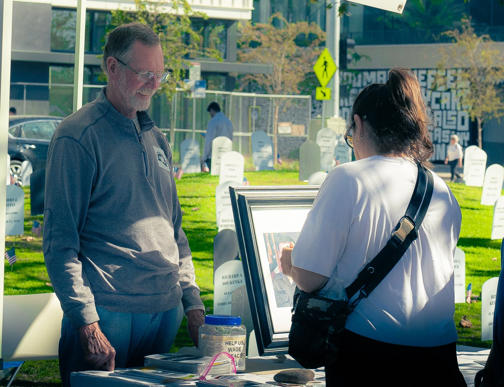 A man and a woman are having a conversation at an outdoor event, with a small table, posters, and a sign that reads "Help Us Wage Peace" visible in the background.