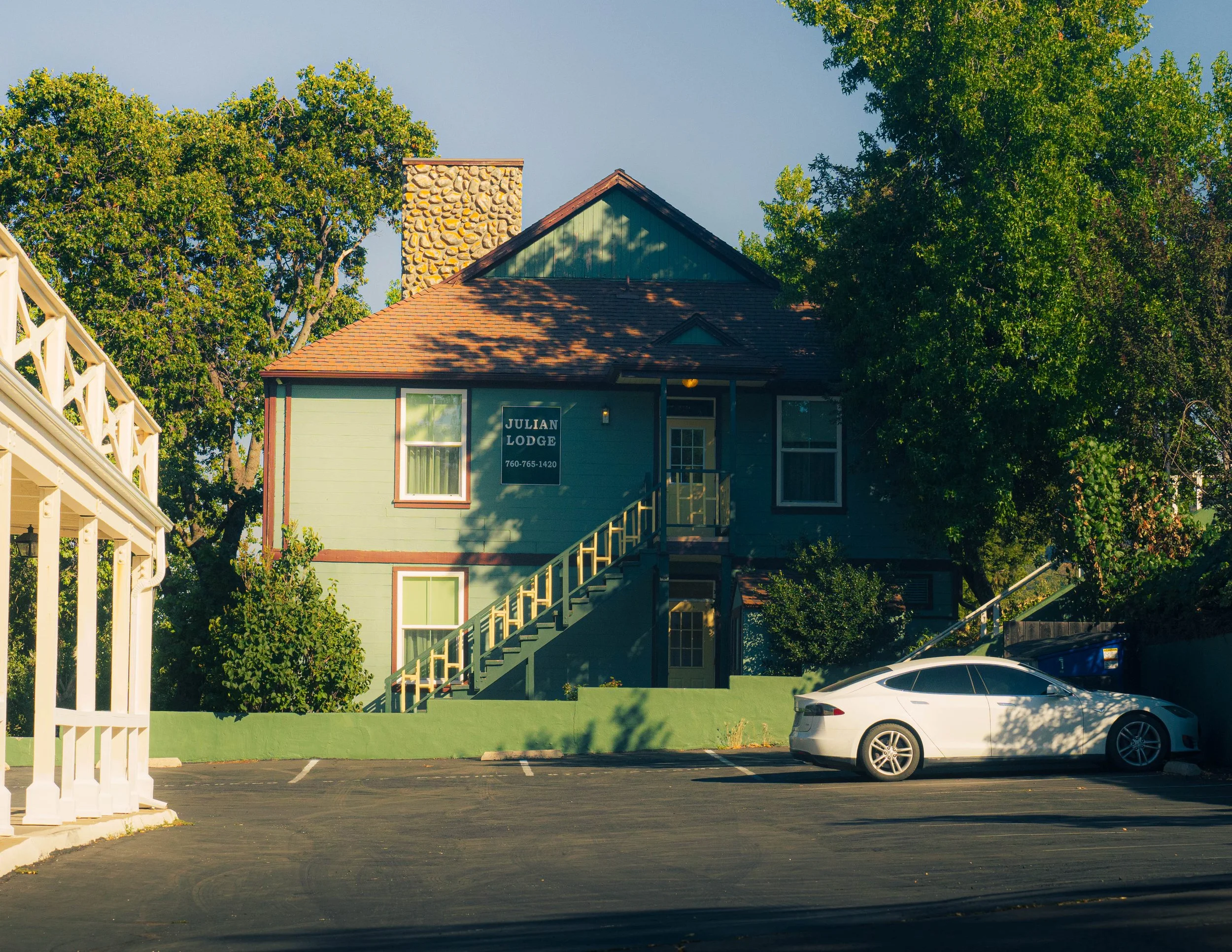 A two-story apartment building with a sign that reads 'Julian Lodge' and a phone number. The building has green siding, a brown roof, and a stone chimney. There are trees around the building, parked cars in the parking lot, and a white wooden fence o