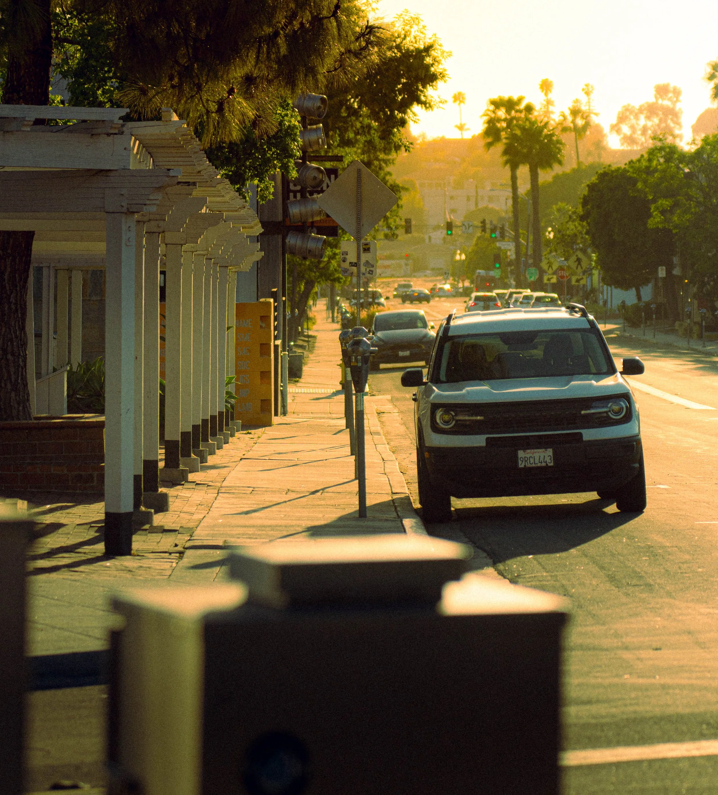 A street scene during sunset with cars parked along the curb, trees lining the sidewalk, and a stop sign in view. The photo is taken from behind a horizontal object that partially blocks the view, possibly a blind or fence.