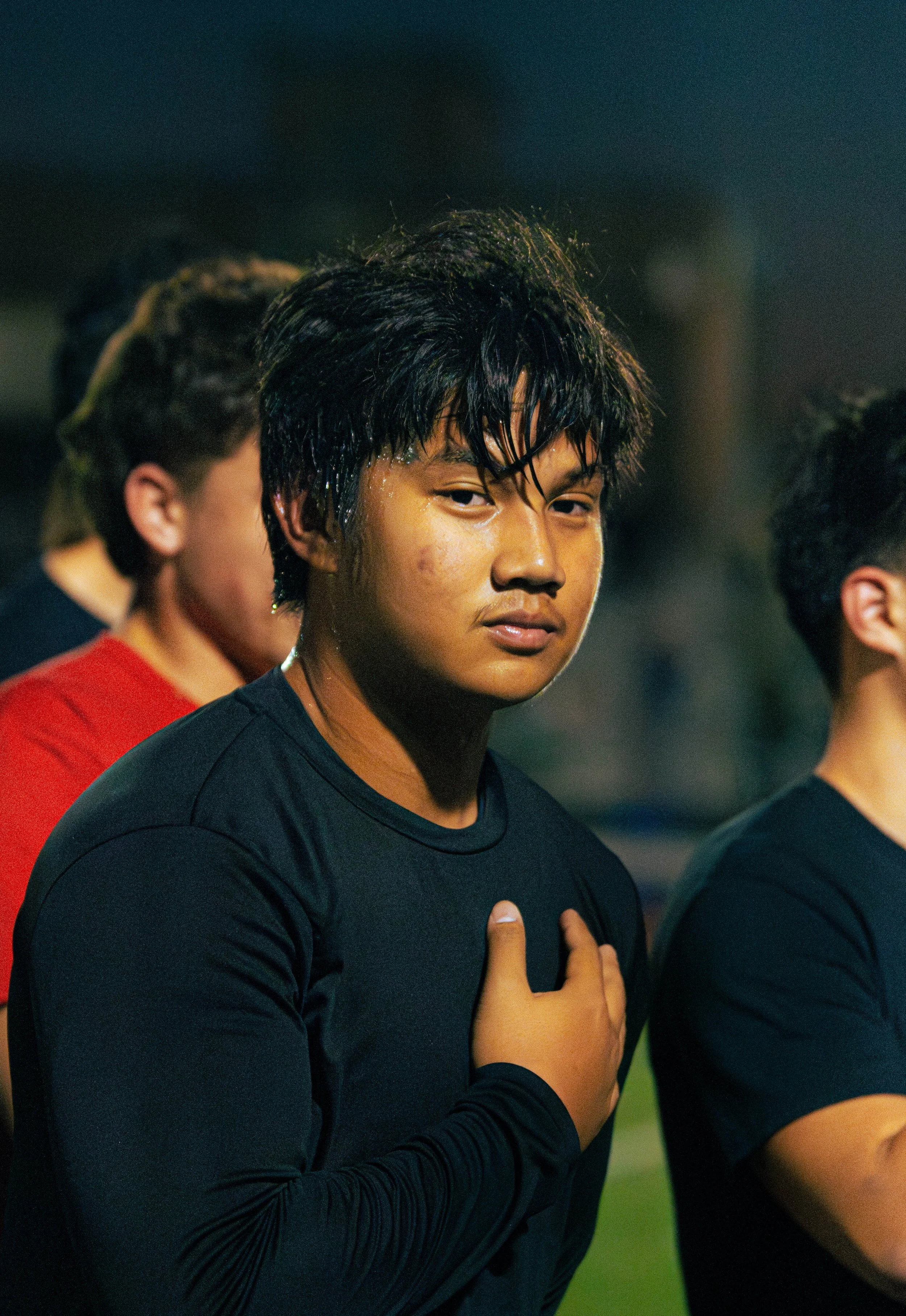 A young person with wet black hair, wearing a black athletic shirt, standing with hand on chest, and closed eyes, likely during a sports practice or game.