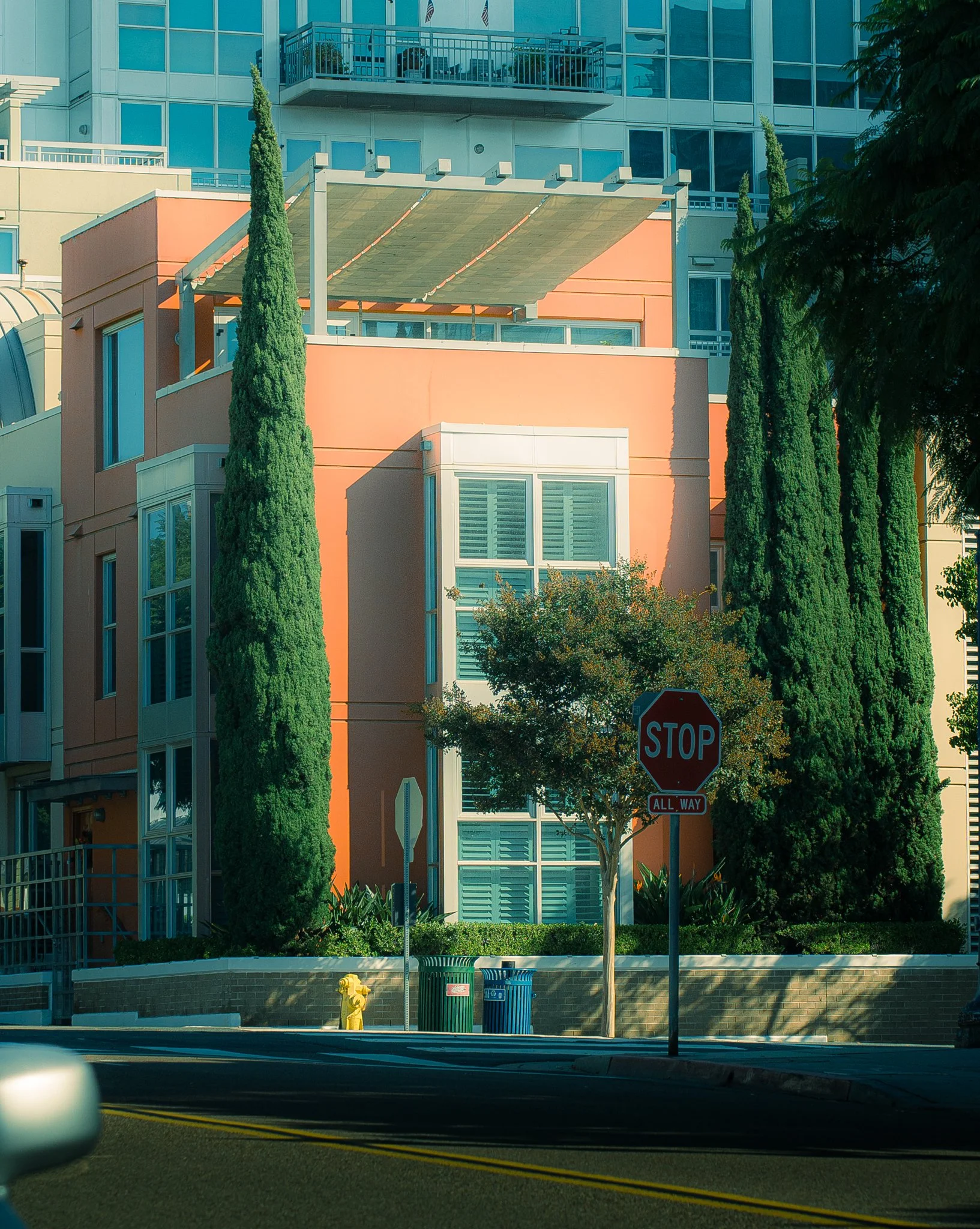 City street scene with modern multi-story building, green trees, a stop sign, trash cans, and a fire hydrant in the foreground.