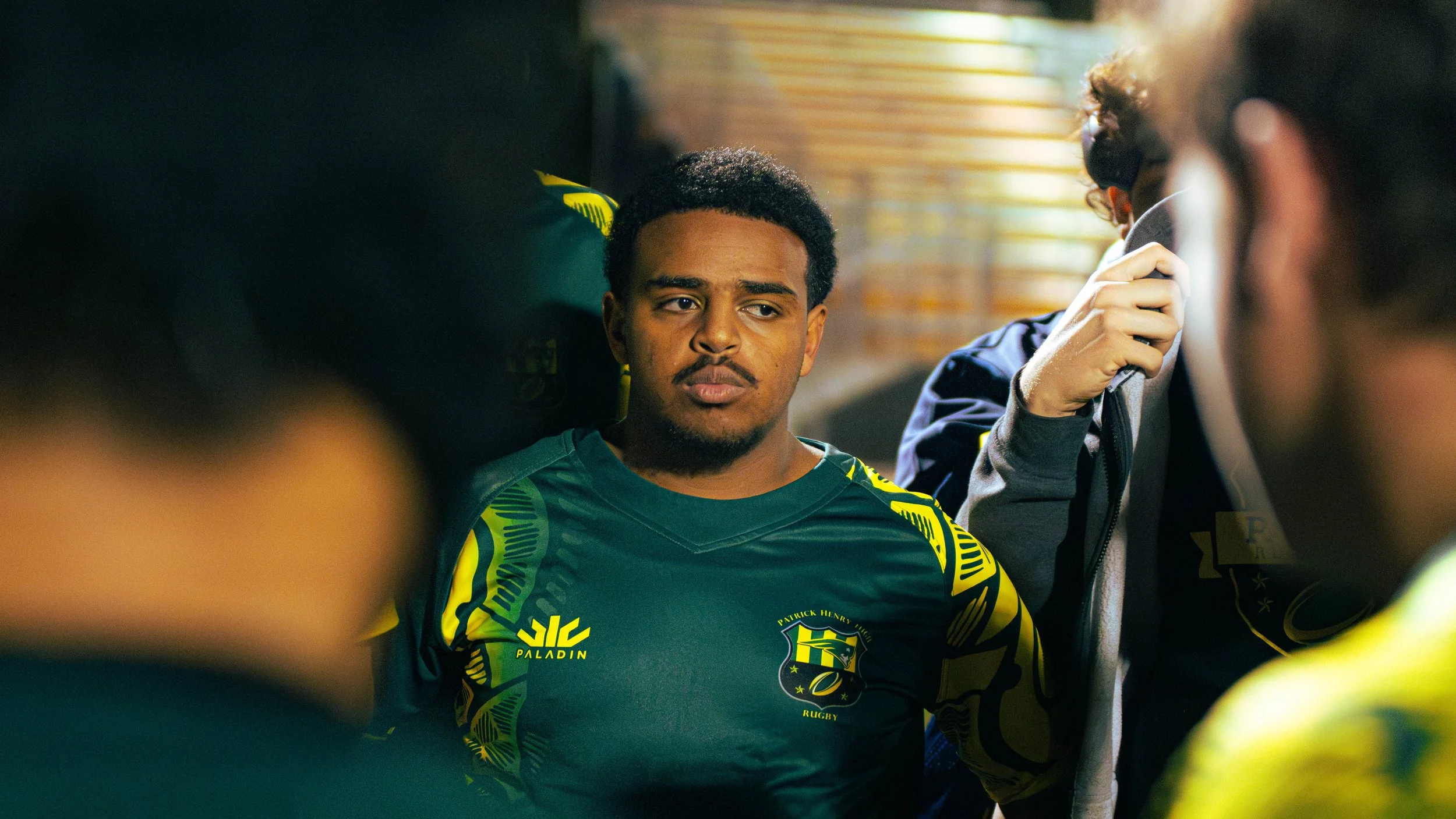 A young man in a rugby uniform sitting with serious expression, surrounded by teammates in a locker room.
