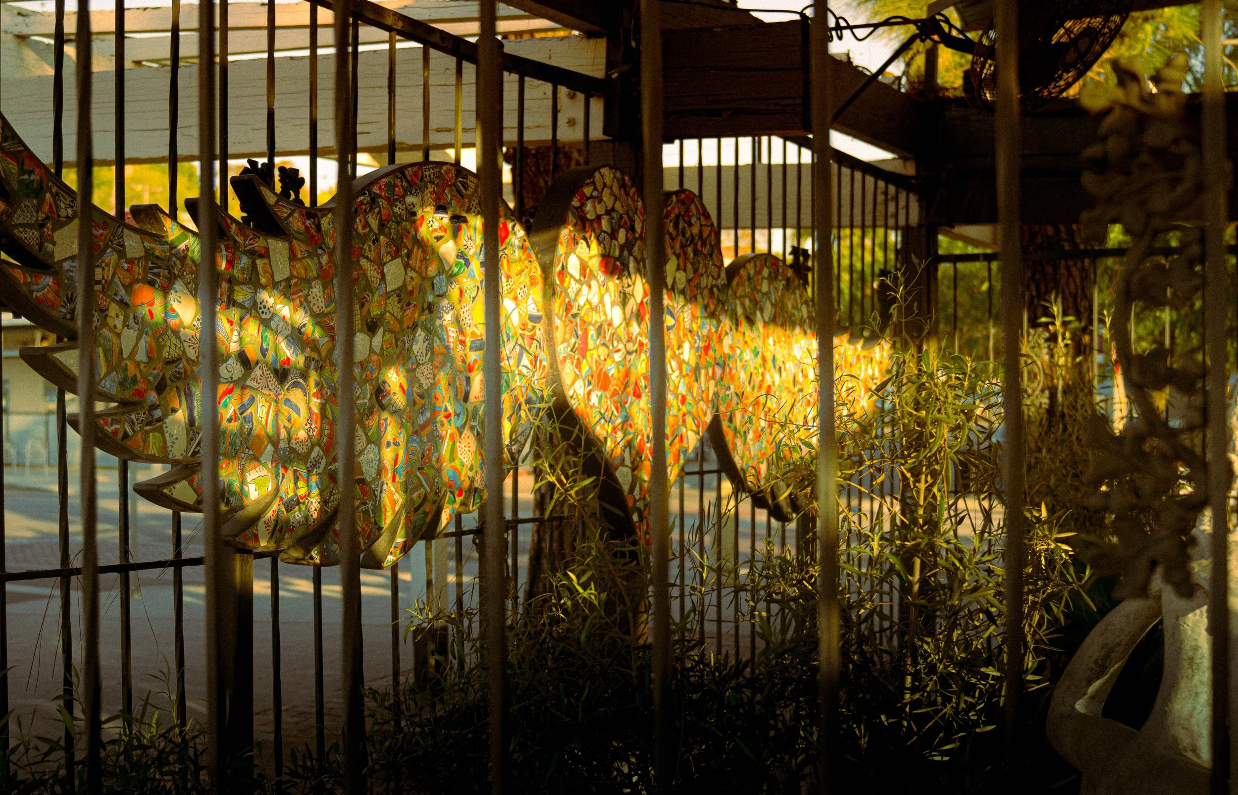 Colorful decorative wooden cutouts hanging on a metal railing, with sunlight shining through them, and greenery visible in the background.