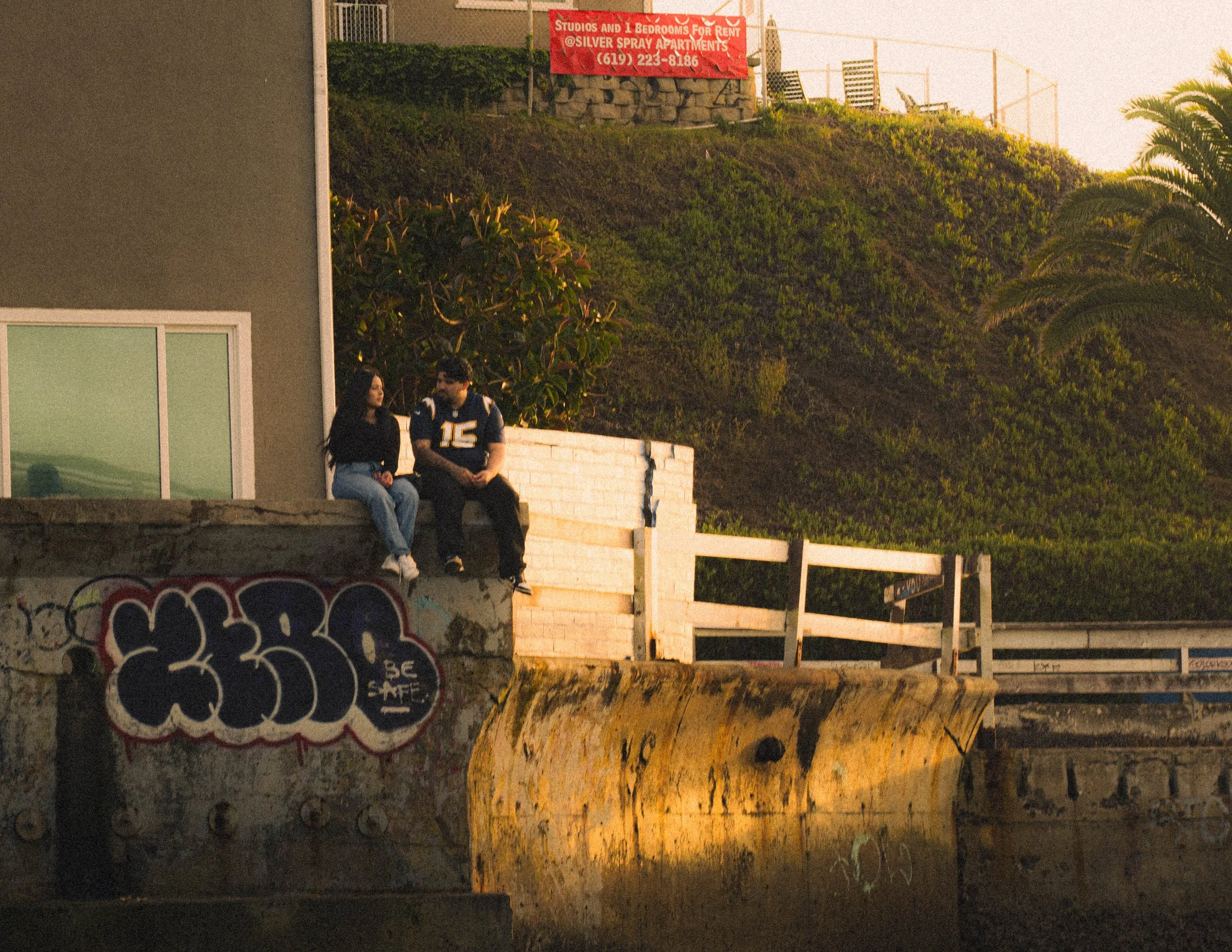 Two people sitting on a ledge outdoors, a woman with long dark hair and jeans and a man wearing a football jersey, at sunset with a hillside and a red sign indicating apartments for rent in the background.