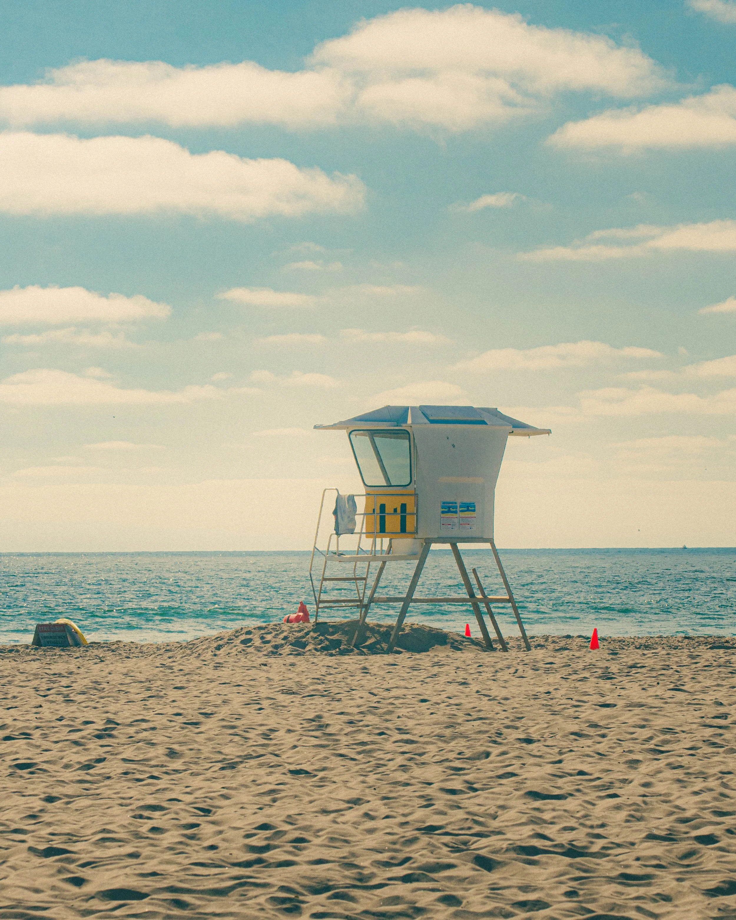 Empty lifeguard tower on a sandy beach with ocean and blue sky with scattered clouds in the background.