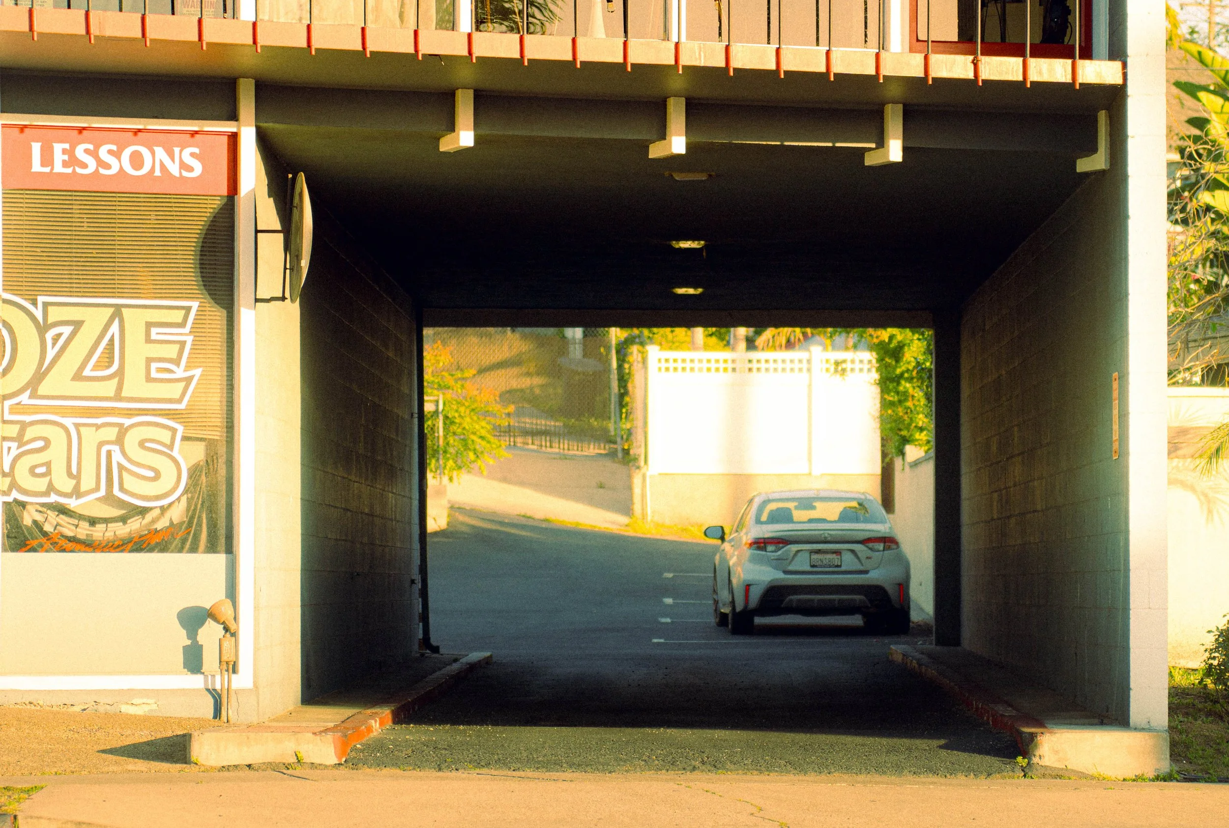 View of a parking garage entrance under an overpass with a silver car driving through. The left side has a sign that says 'LESSONS' and a partial view of an advertisement with the words 'OZE' visible. The background shows trees and fencing, and the t
