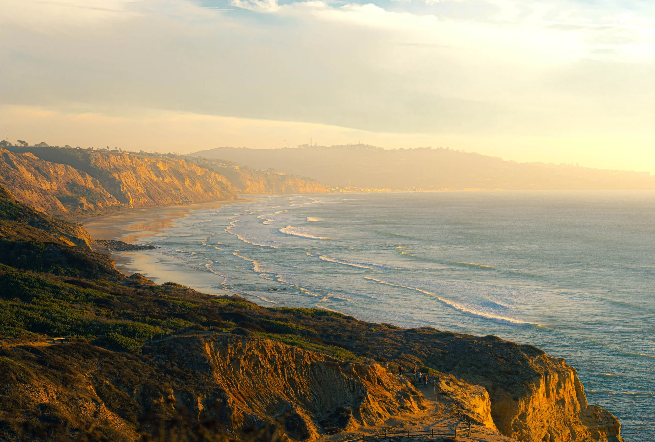 Sunset over rugged coastline with cliffs and waves crashing onto the beach, with a few people walking along a path on the cliffs.