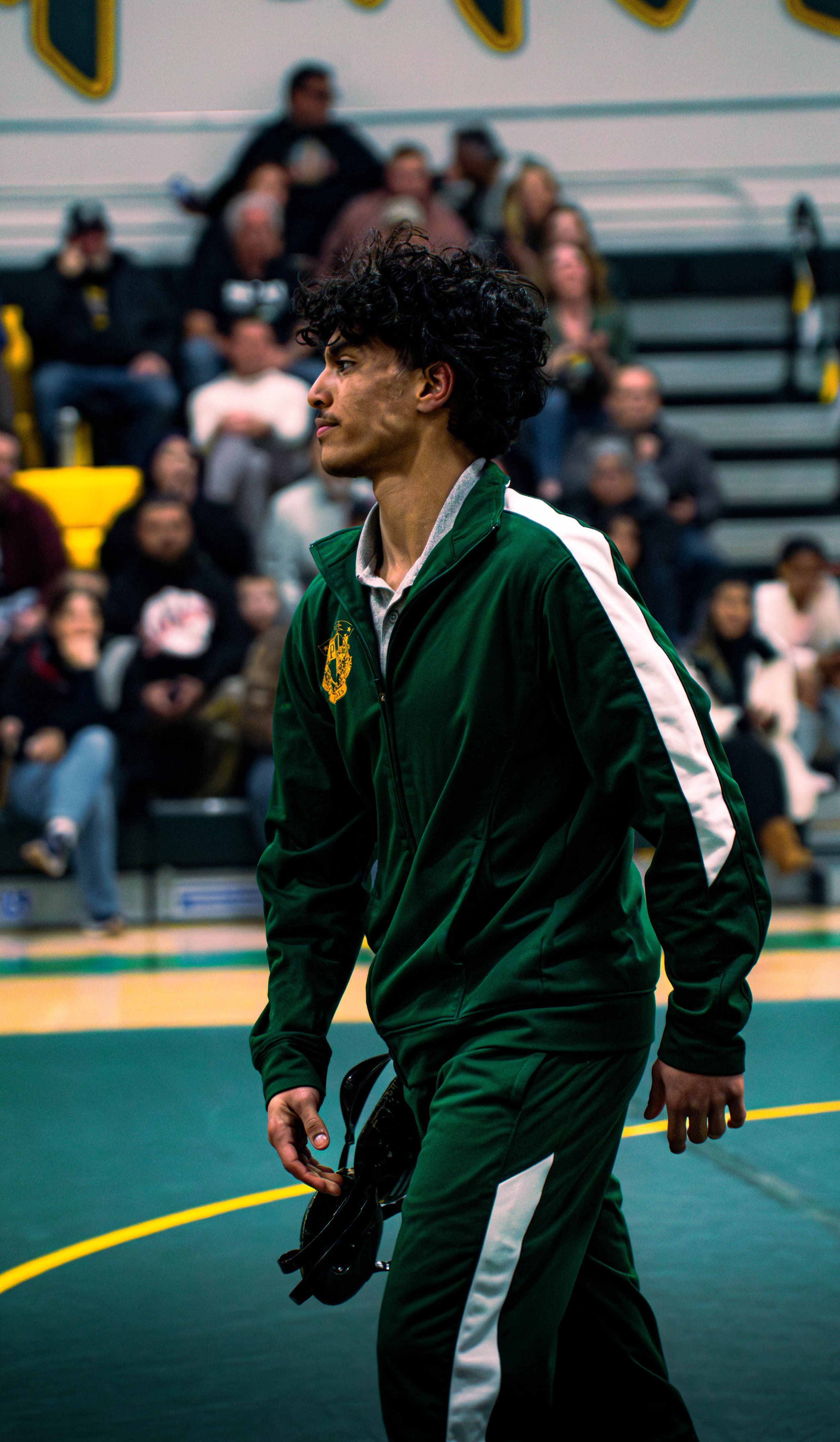 A man with curly black hair wearing a green and white sports jacket, walking on the gym floor holding a pair of black shoes in his right hand, with people seated on bleachers in the background.