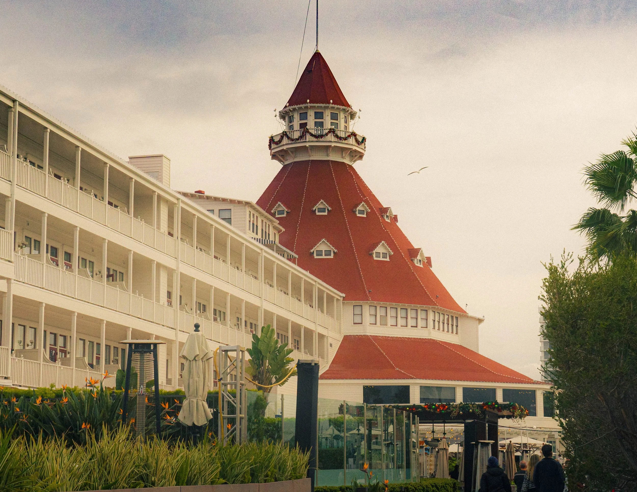 A building with white railings and balconies on the left side of the image. In the center, there is a tall, red-roofed tower with small windows and a decorative balcony, topped with a spire. In the foreground, there are green plants and orange flower