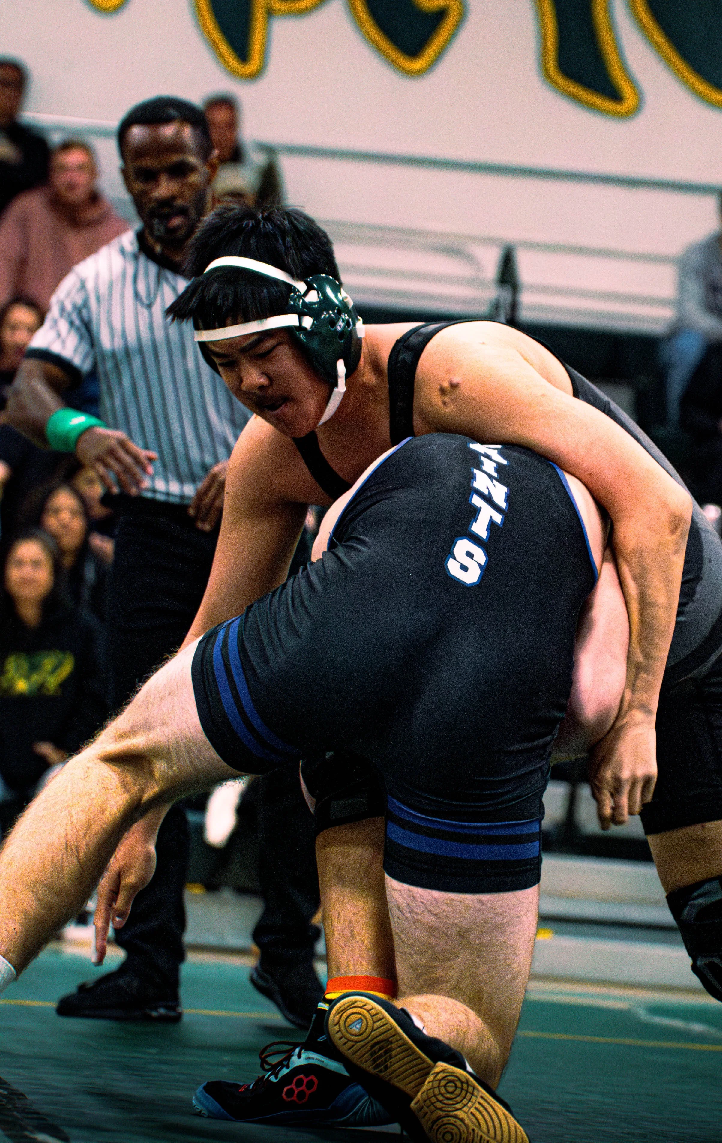 Two male wrestlers in a wrestling match on a mat, with a referee observing in the background and spectators watching.