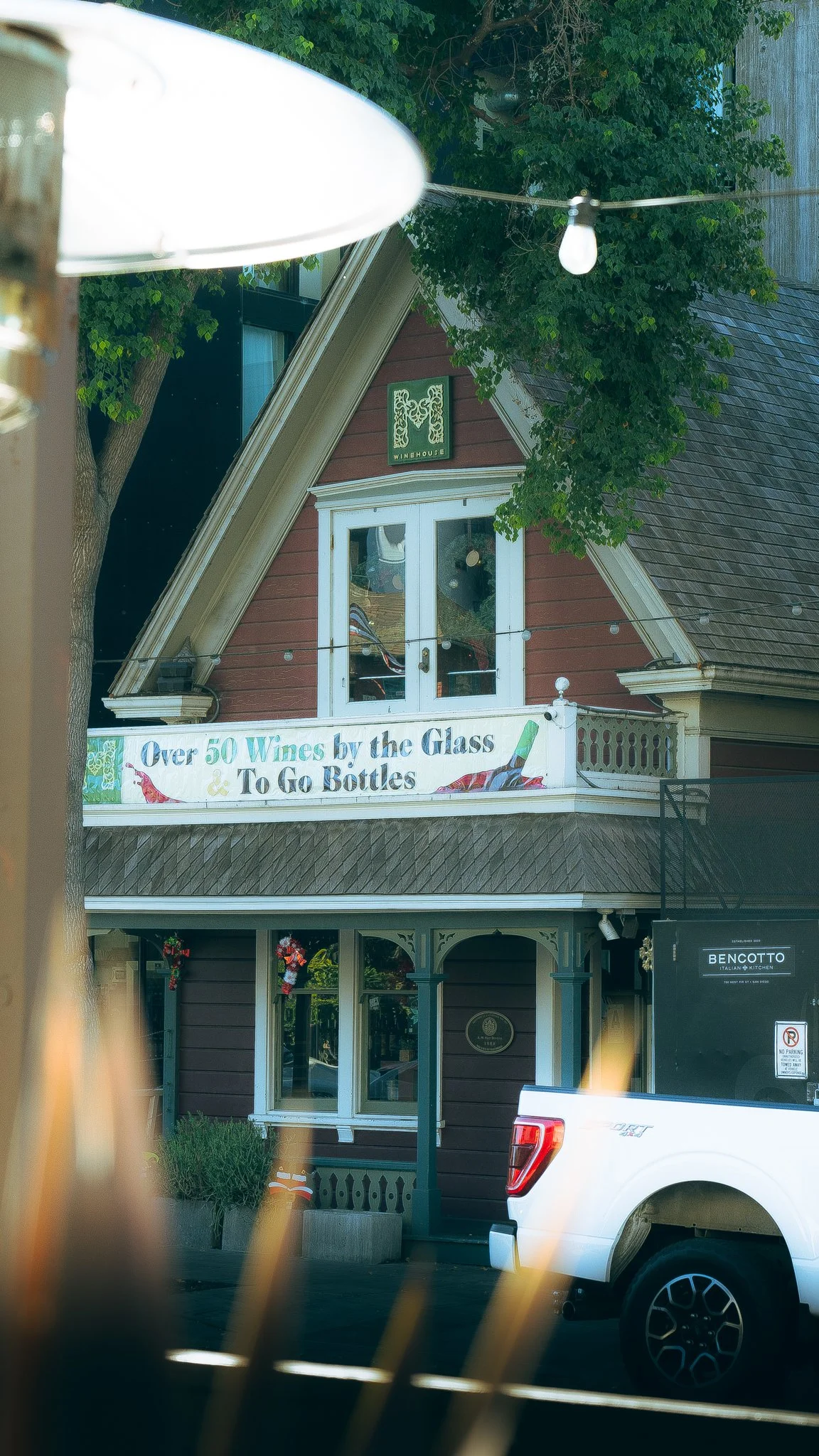 Exterior of a building with a sign advertising over 50 wines by the glass and to go bottles, a tree partially blocking the view, and a white pickup truck parked in front.