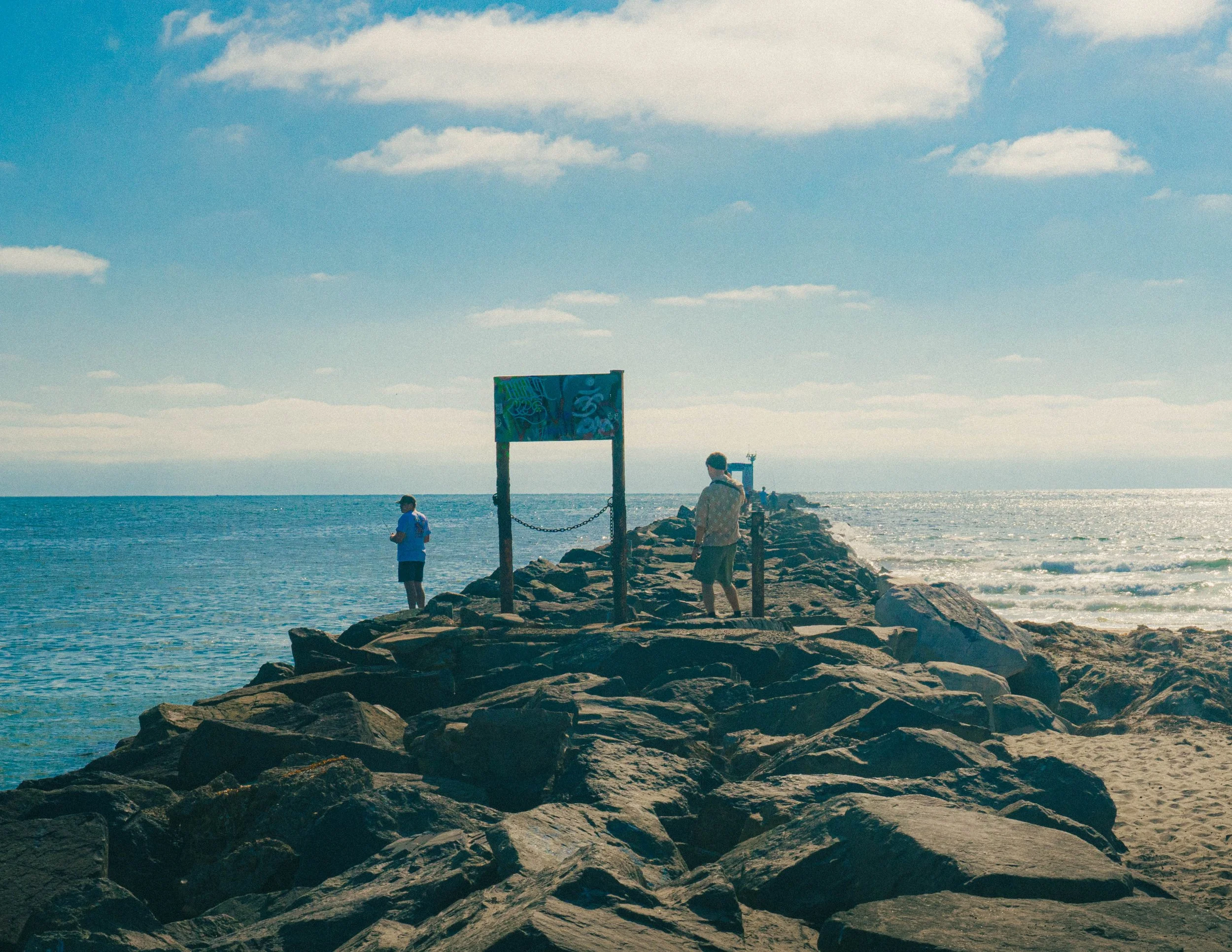 People on a rocky pier at the beach with a colorful sign, ocean waves, and a partly cloudy sky.
