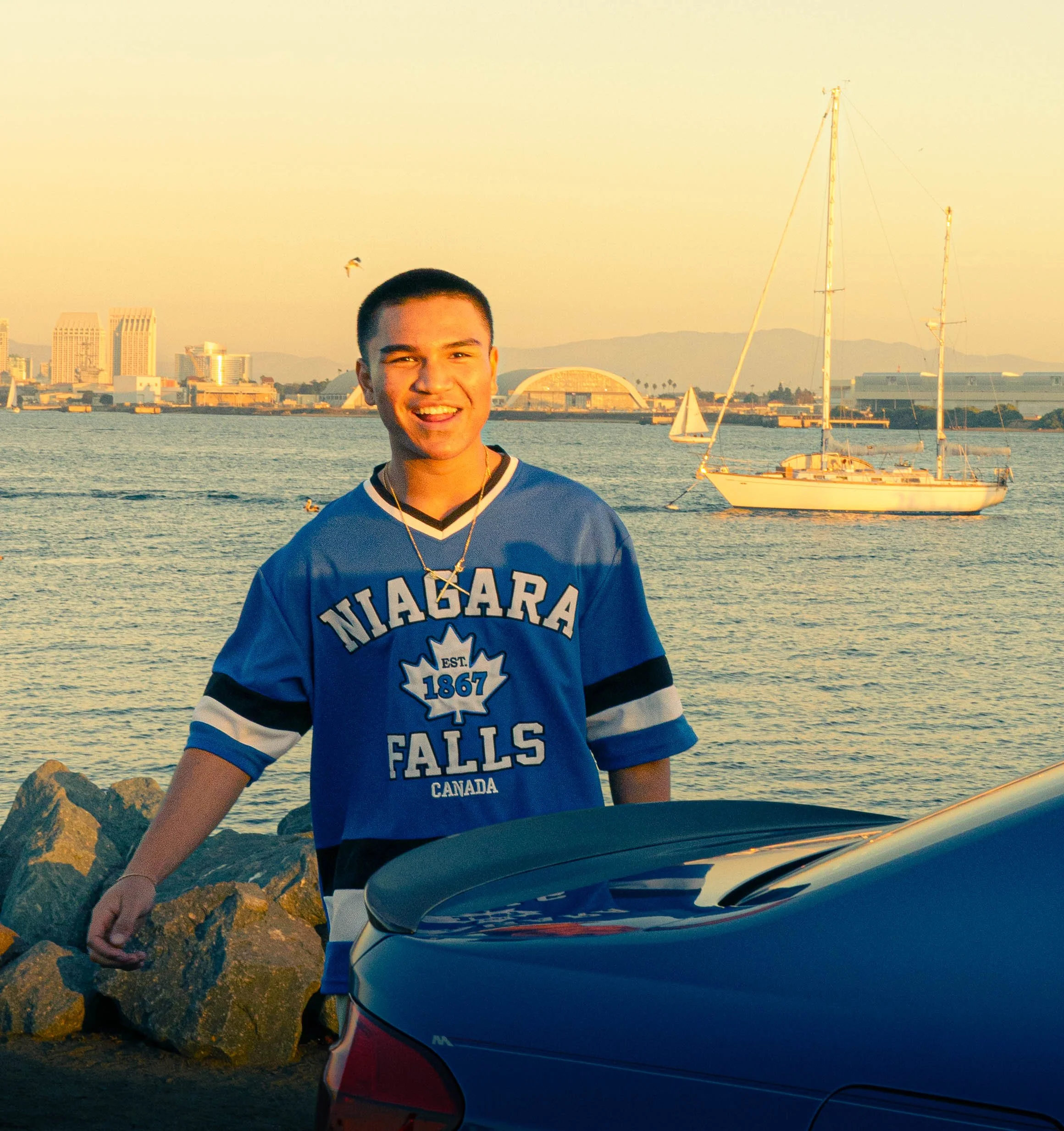 A young man smiling at the camera standing outdoors near a harbor with sailing boats and city skyline in the background during sunset.