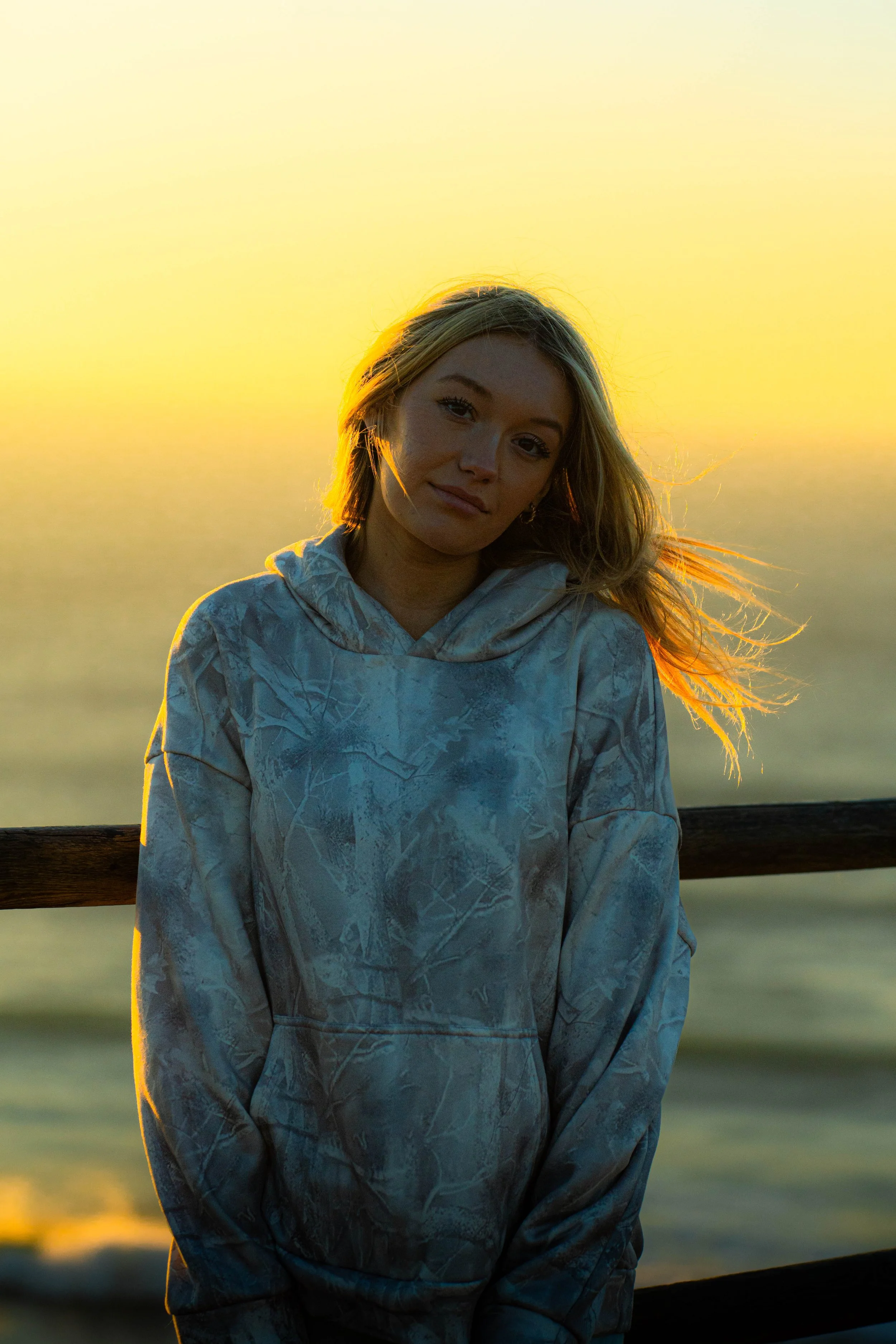 A young woman with shoulder-length blonde hair standing outdoors at sunset, wearing a camouflage-patterned hoodie, with a calm expression and a blurred background of water and a yellow-orange sky.