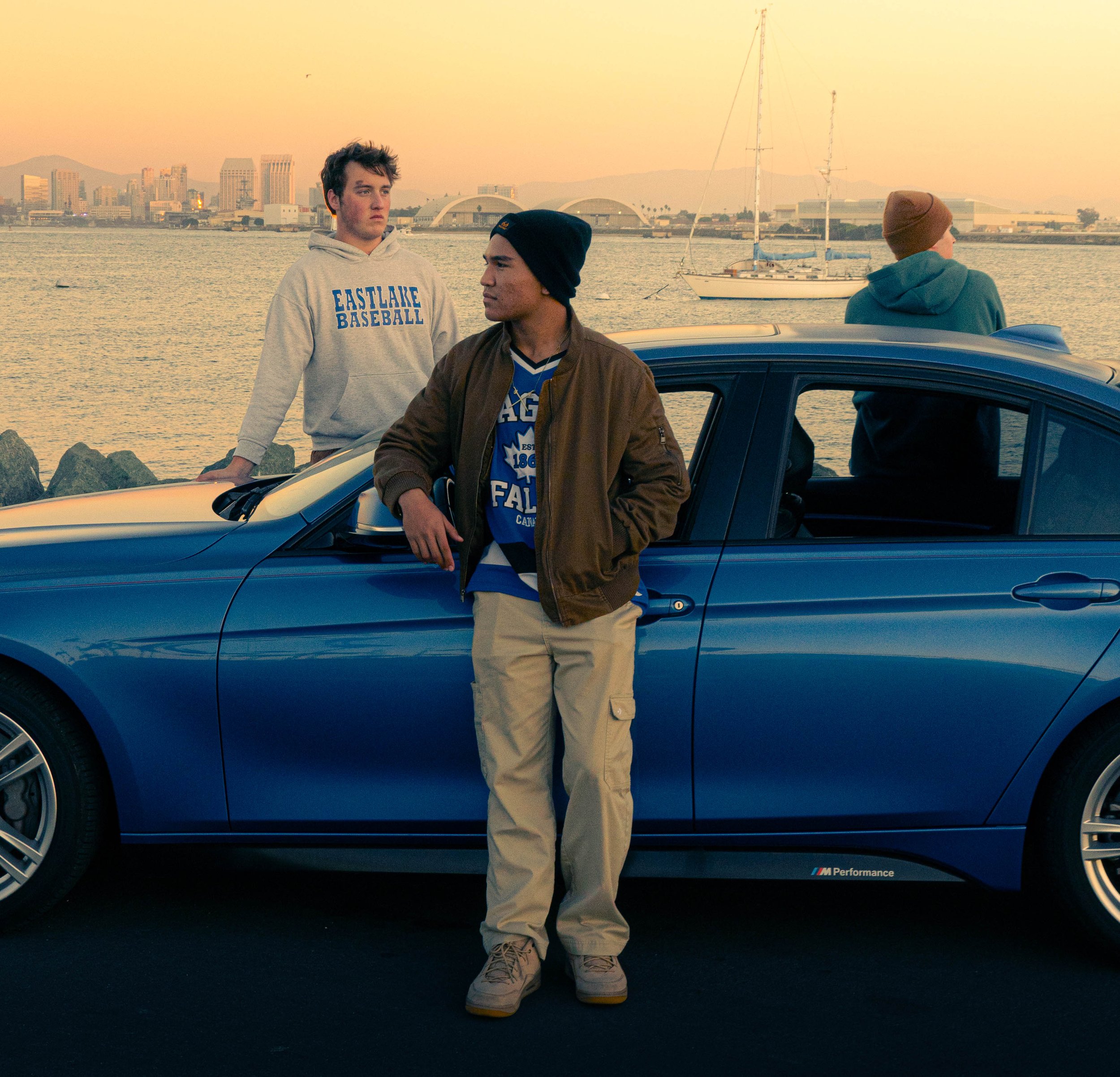 Three young men near a blue car with a city skyline, sailboats, and mountains in the background during sunset. One is standing and wearing a beige hoodie, another leaning on the car in a brown jacket and beanie, and the third facing away wearing a bl