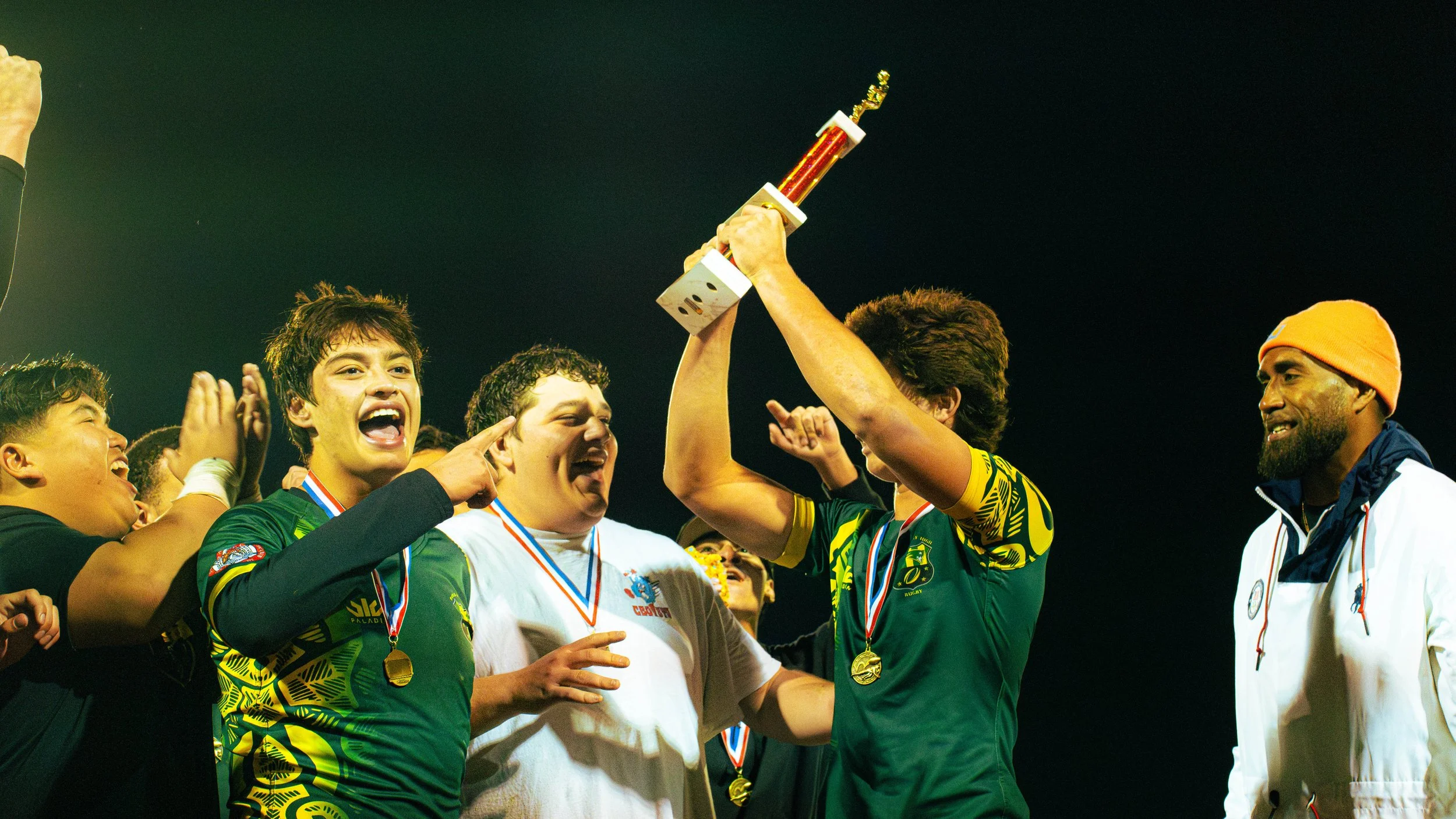 A group of athletes celebrating on a sports field, holding a trophy, and wearing medals. They are smiling and cheering, with some pointing at the trophy.