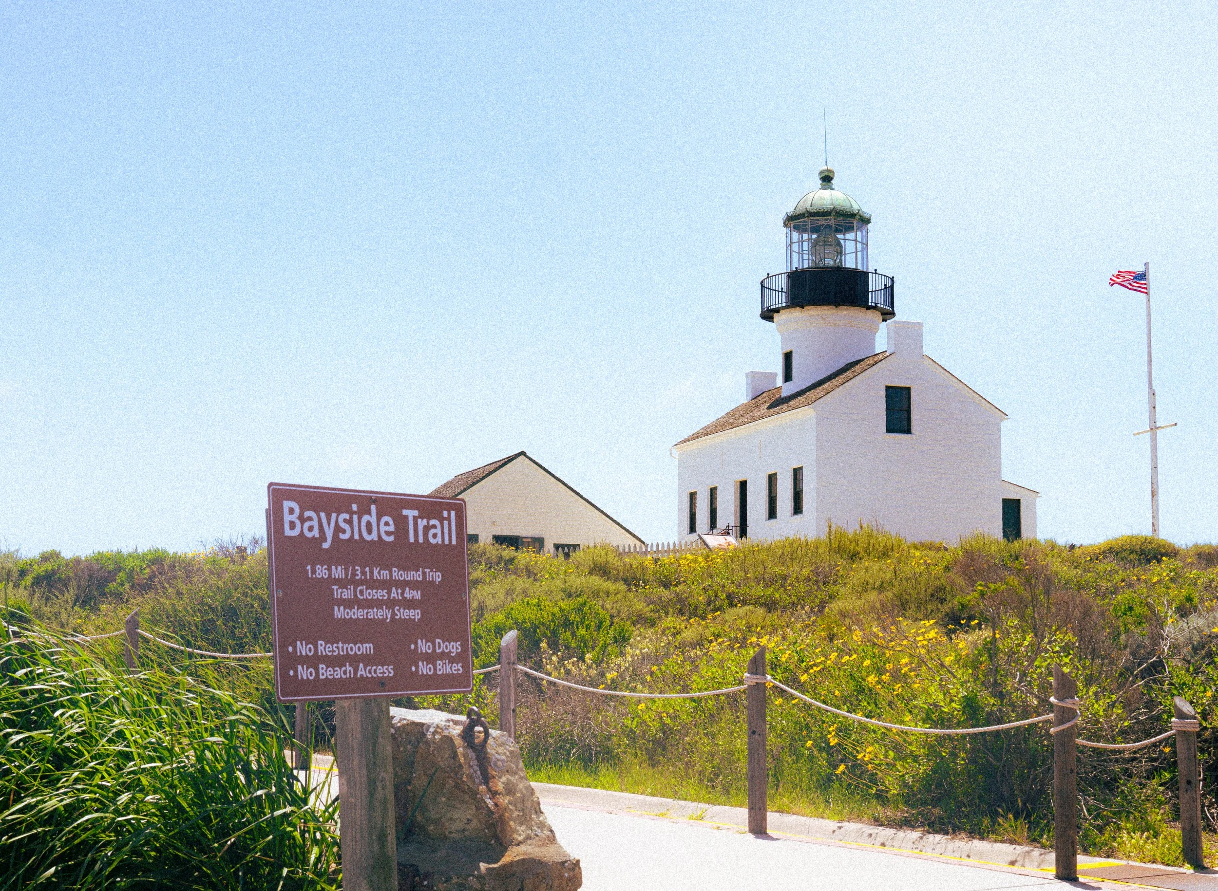 A white lighthouse with black railing on top, a flagpole with an American flag, and a brown trail sign with white text in the foreground, surrounded by green bushes and grass, under a clear blue sky.