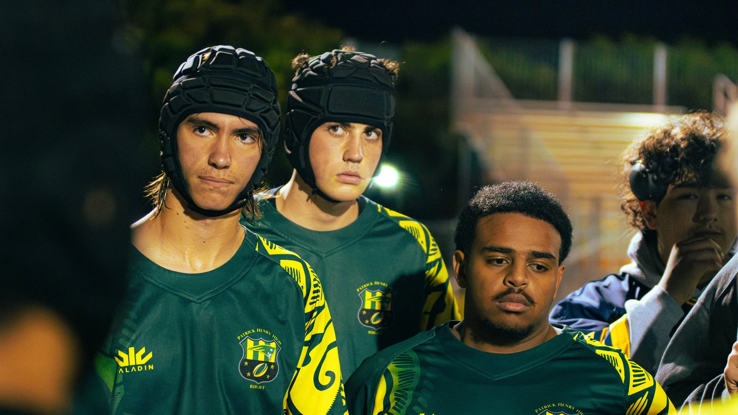 Group of young male athletes wearing green and yellow sports jerseys and black protective headgear, standing closely together during a night-time sports event.