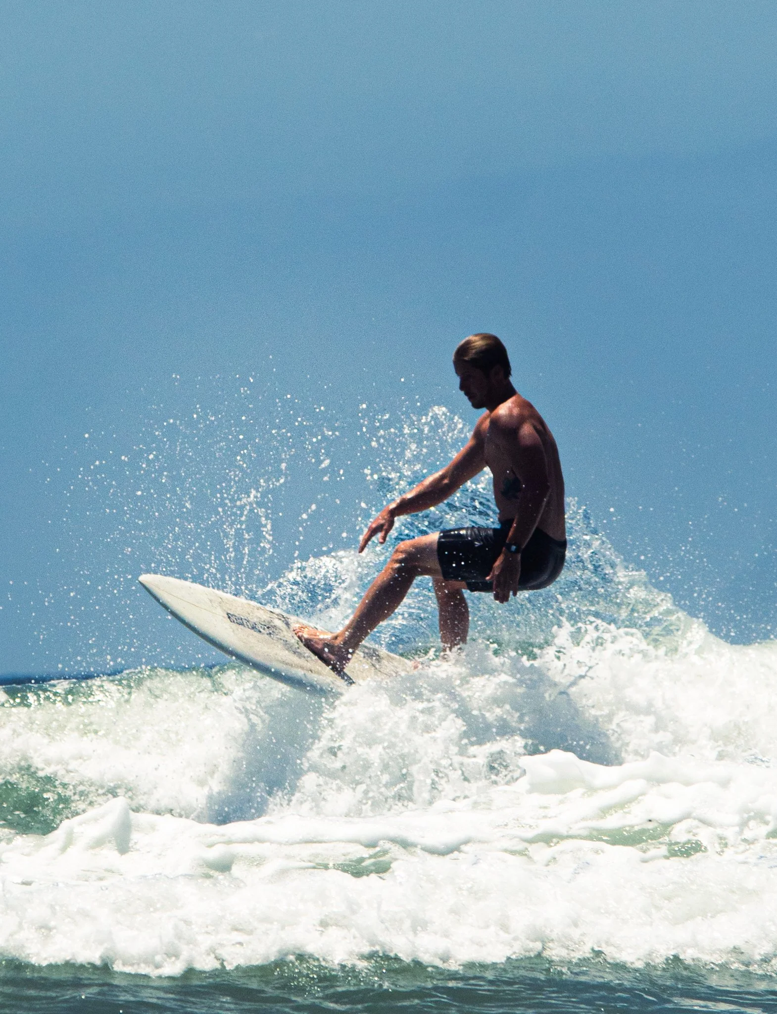 A young man with tan skin and brown hair surfing on a wave in the ocean, wearing black swim trunks.