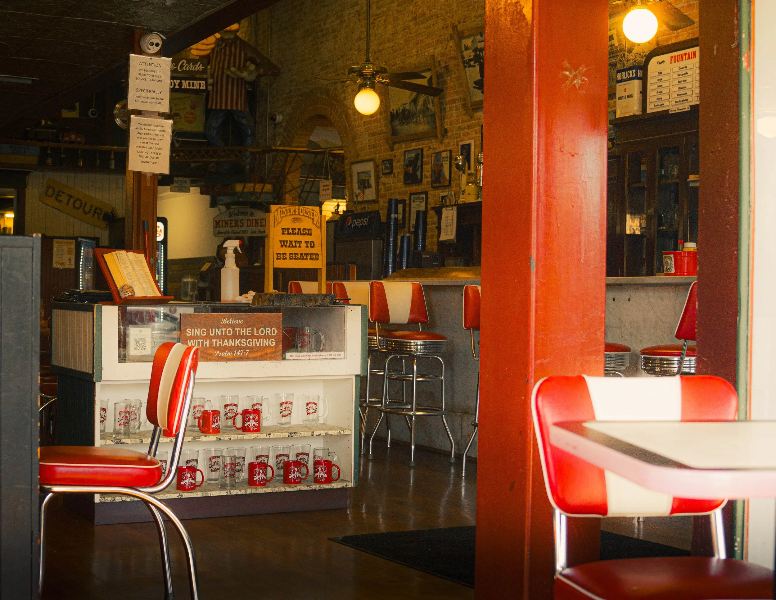 Interior of a vintage-style diner with red and white chairs, a sign that reads 'Please wait to be seated,' and shelves with red mugs and glasses. There are framed pictures on the brick walls and a sign with a biblical verse about Thanksgiving.