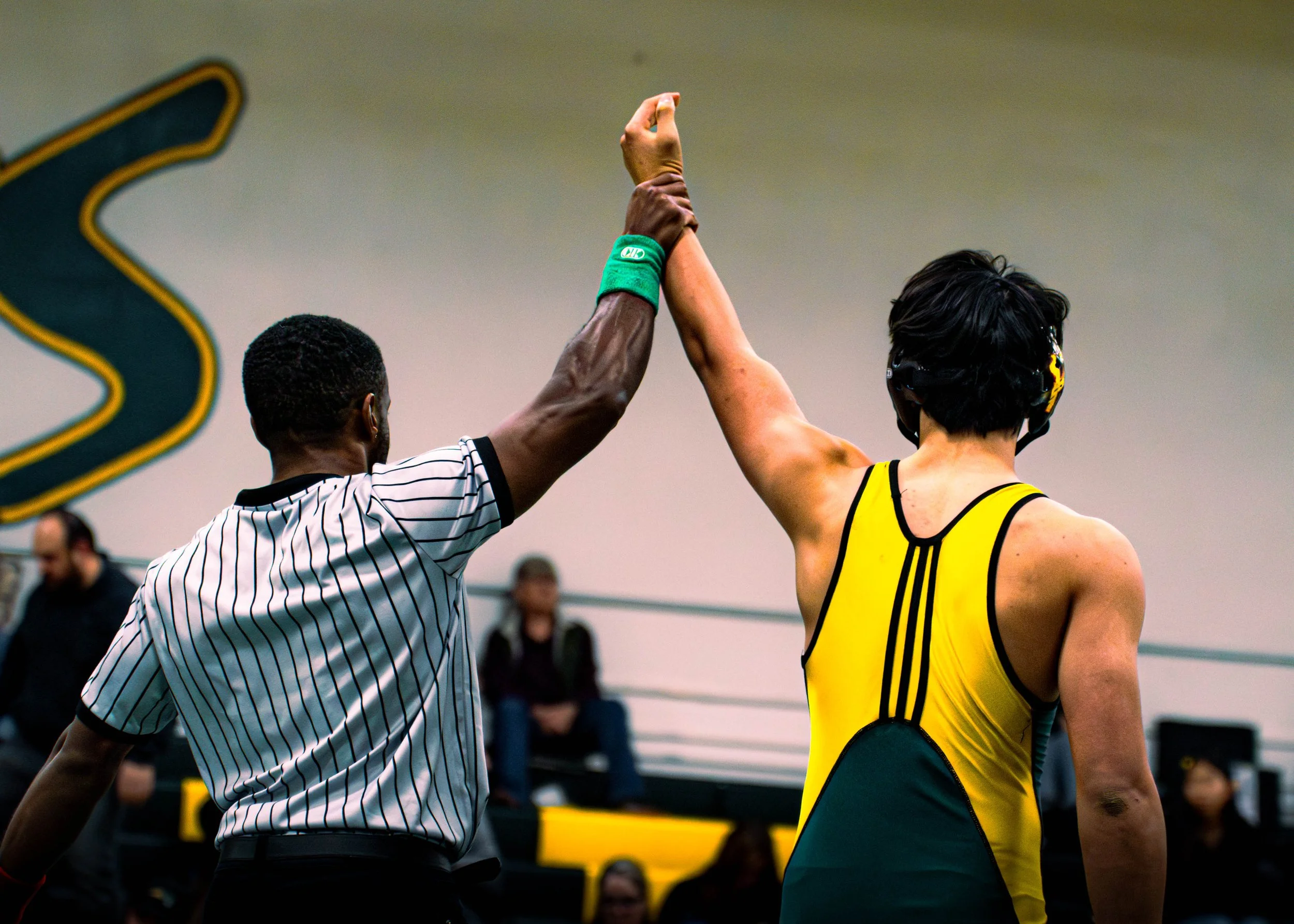 A referee with dark hair and a striped shirt high fives a wrestler with short black hair, wearing a yellow and green singlet, inside an athletic facility with an audience and scoreboard in the background.