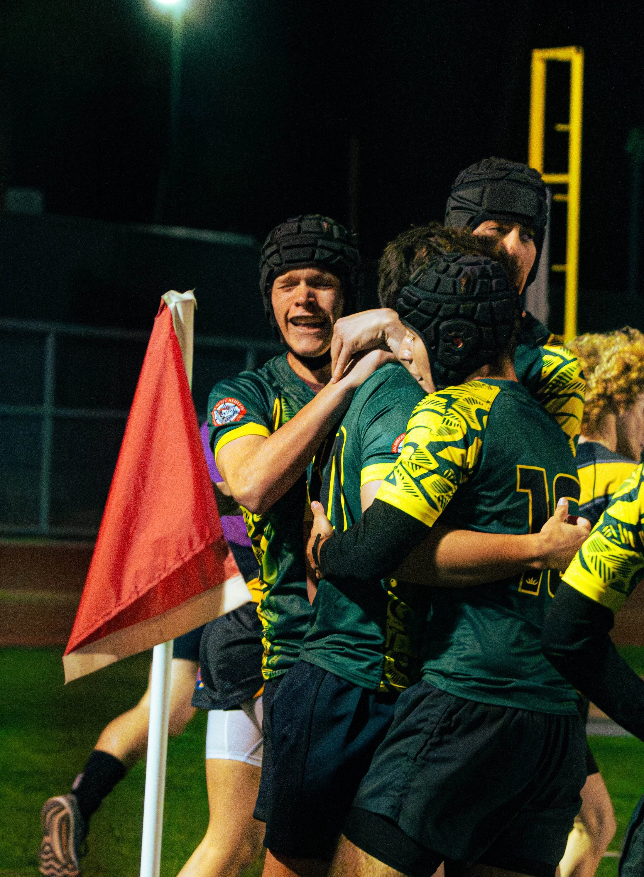 Rugby players celebrating on the field at night, wearing black and yellow uniforms and black helmets.